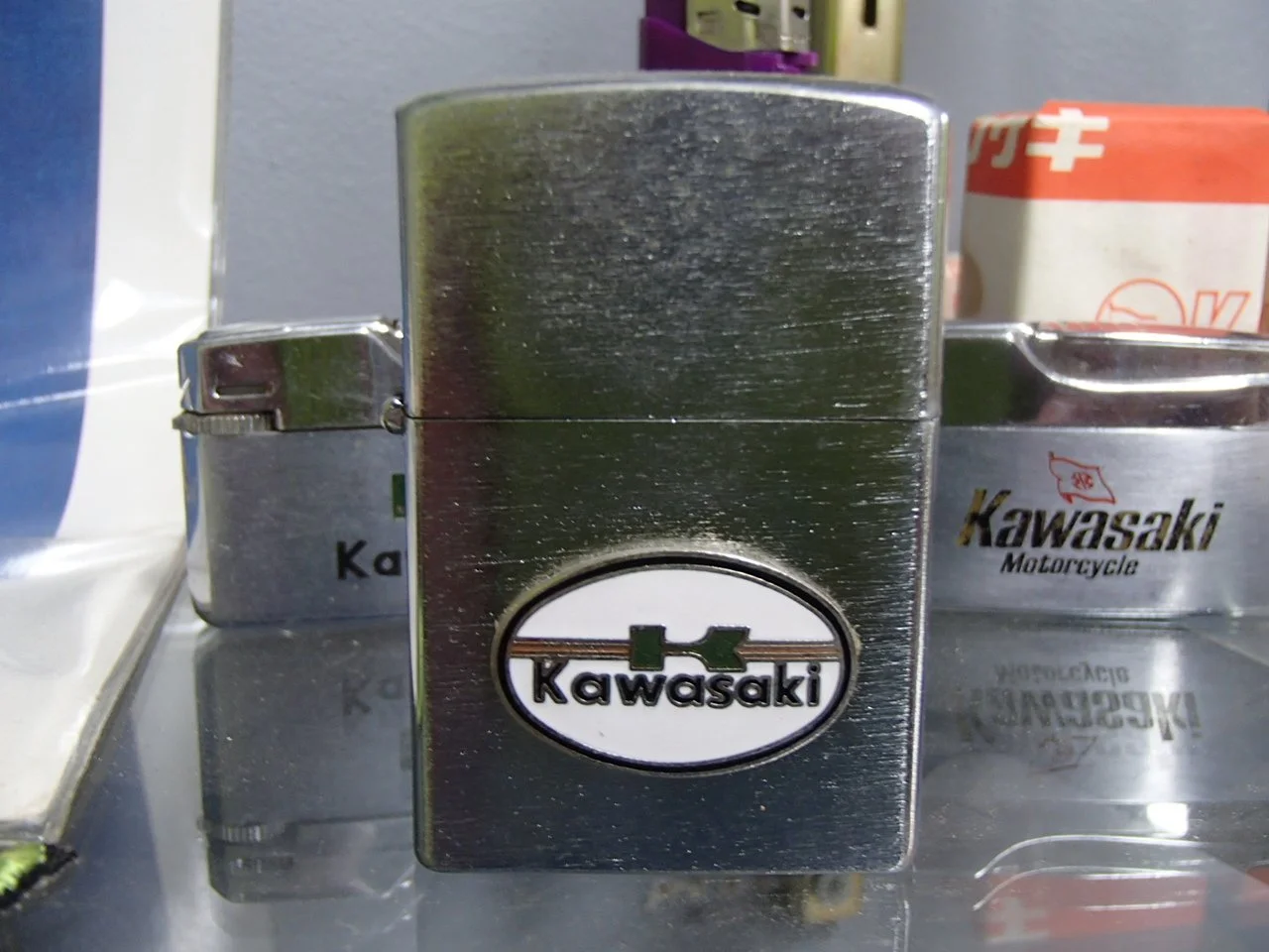 A shiny metal lighter with a Kawasaki logo in the center, placed on a reflective surface with another similar lighter and a box or container in the background.