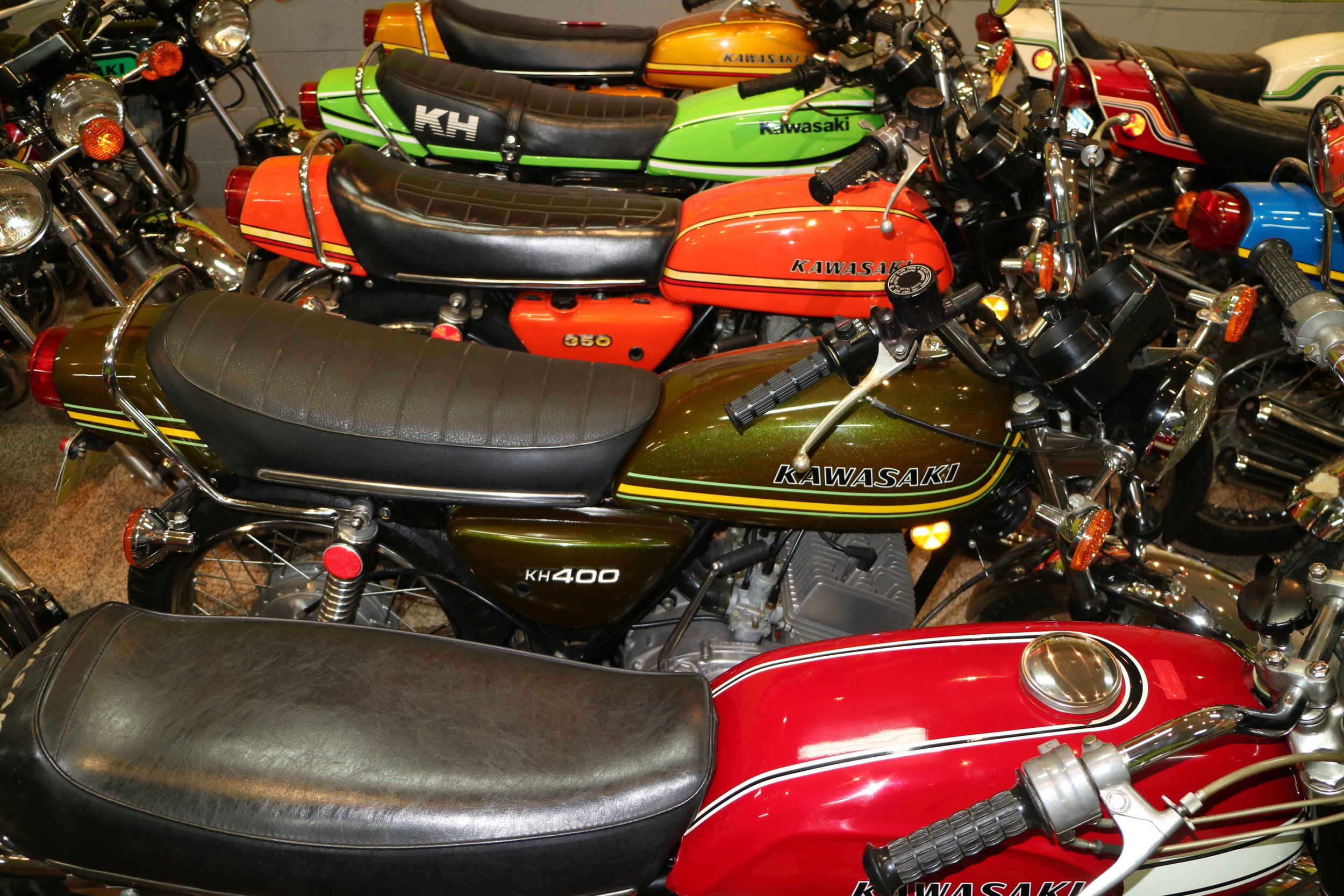 Multiple vintage Kawasaki motorcycles parked closely together, featuring various colors including red, green, orange, blue, and brown.