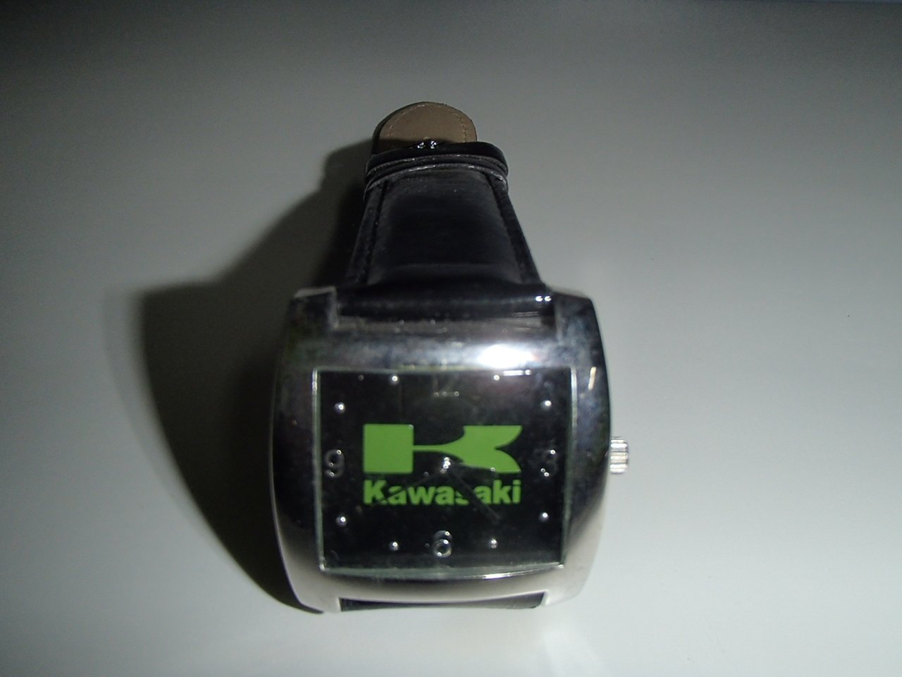 A digital wristwatch with a black face, displaying a green graphic of the number 3, the word 'Kawasaki,' and a black strap.