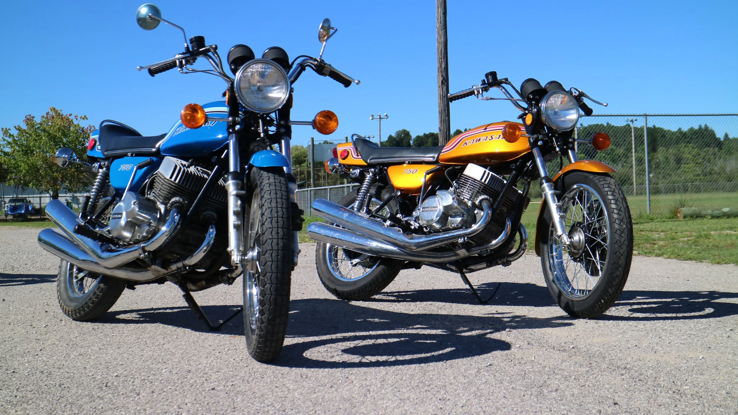 Two vintage motorcycles parked on a gravel surface with a chain-link fence and trees in the background under a clear blue sky.