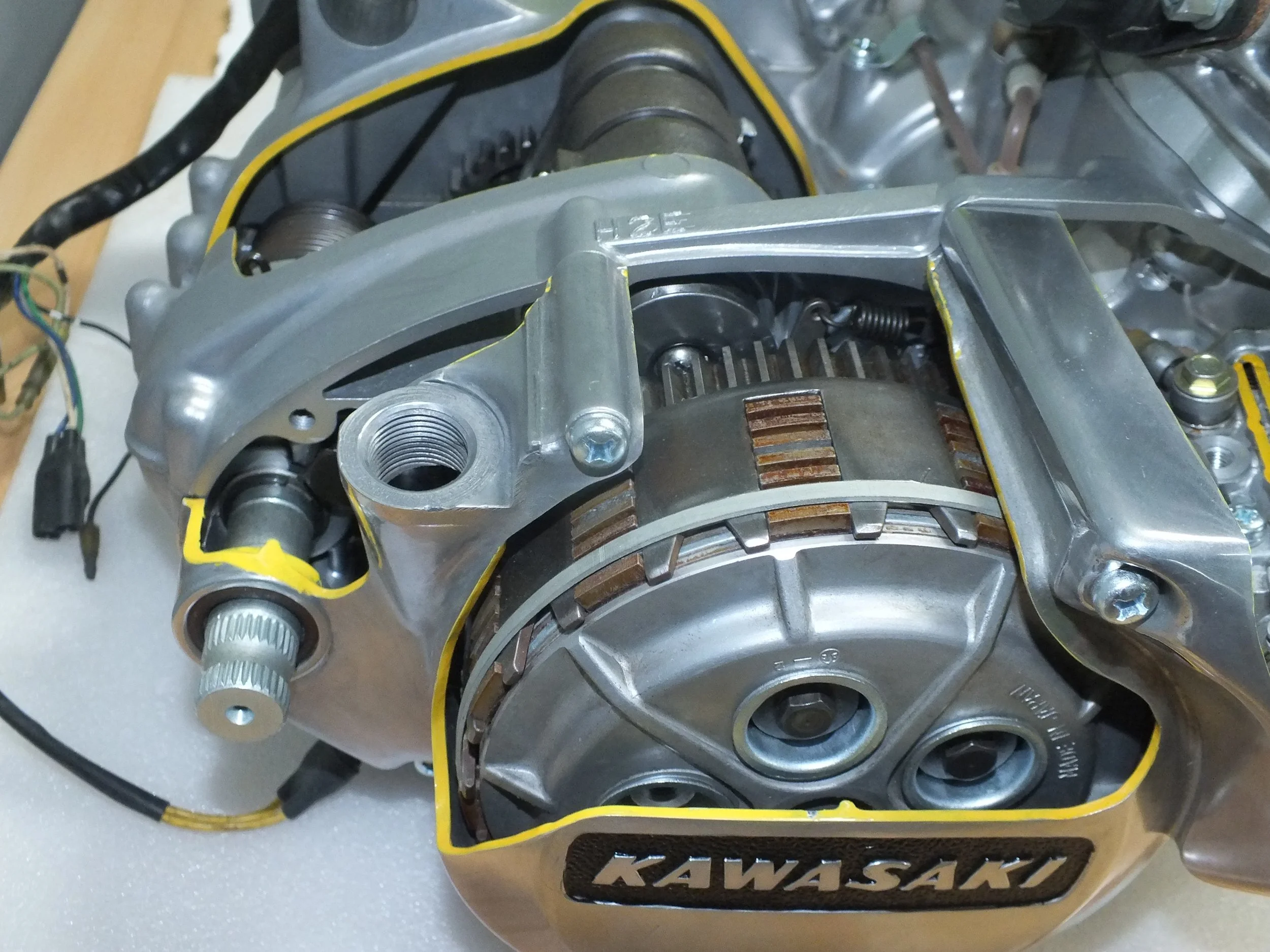 Close-up view of a Kawasaki motorcycle engine part with metal components, gears, and wiring, partially surrounded by a yellow border.
