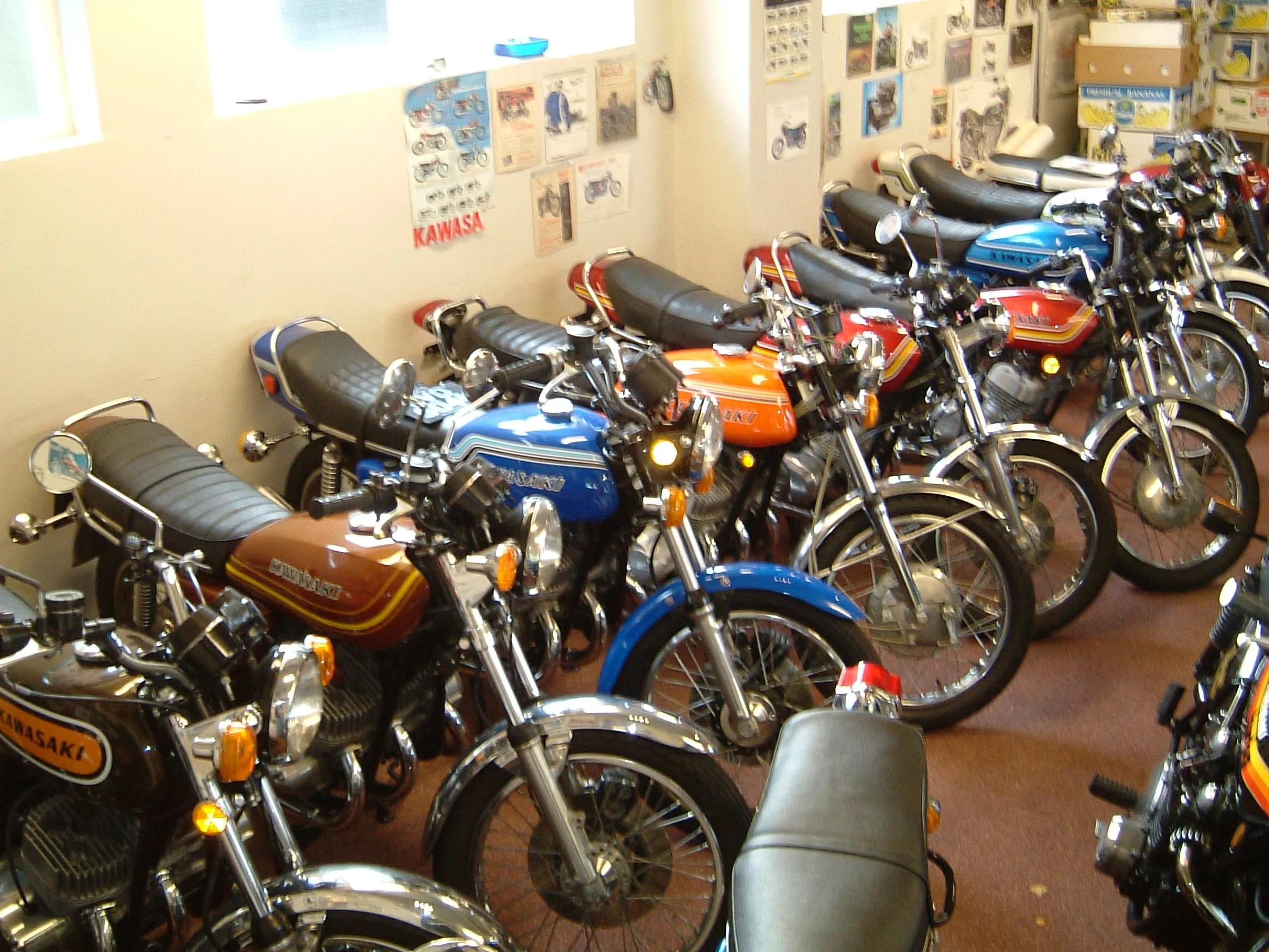 Multiple vintage Kawasaki motorcycles parked in a room with posters and boxes on the wall.