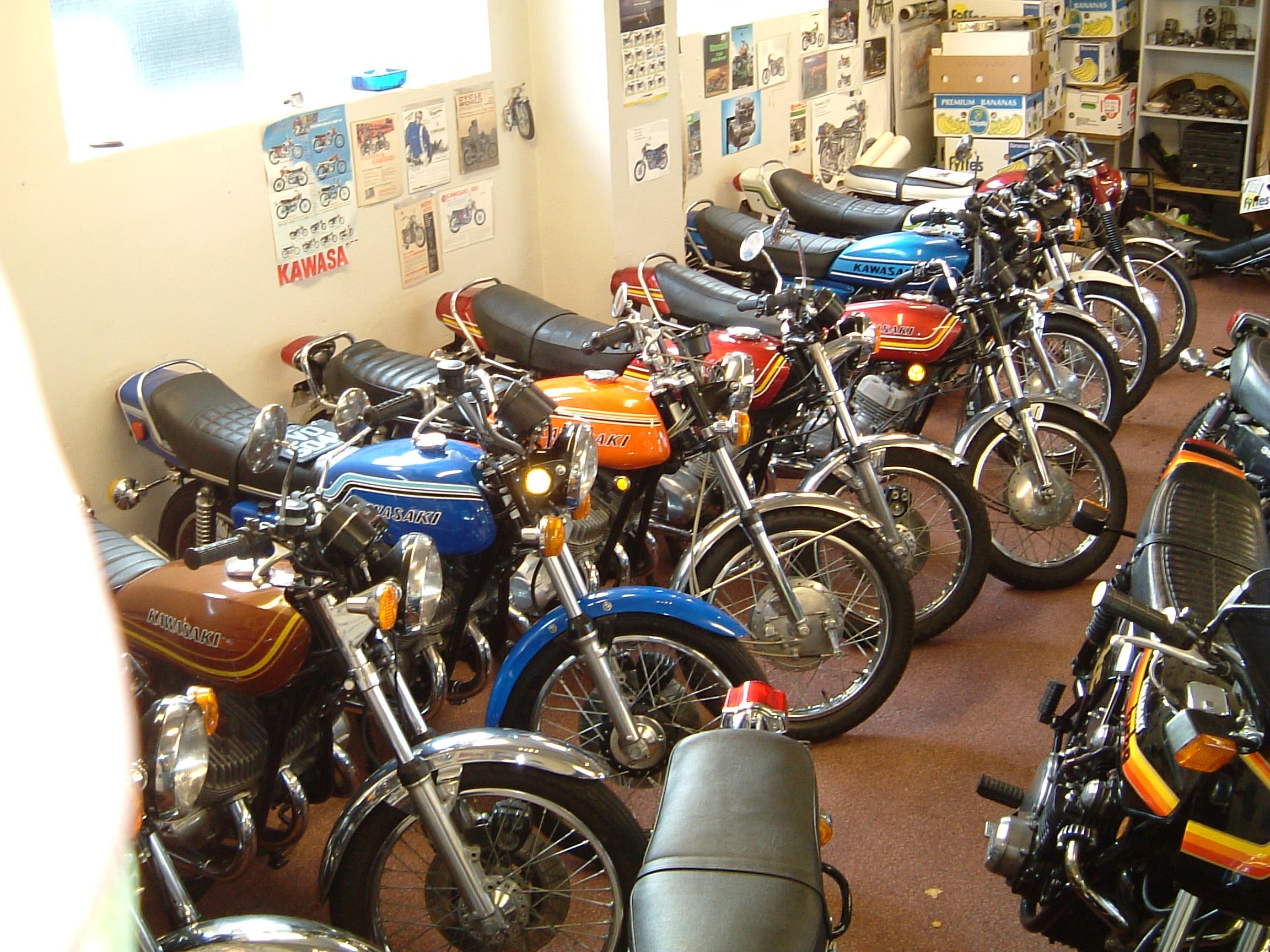 A room filled with vintage Kawasaki motorcycles lined up side by side, with posters and photos of motorcycles decorating the walls.