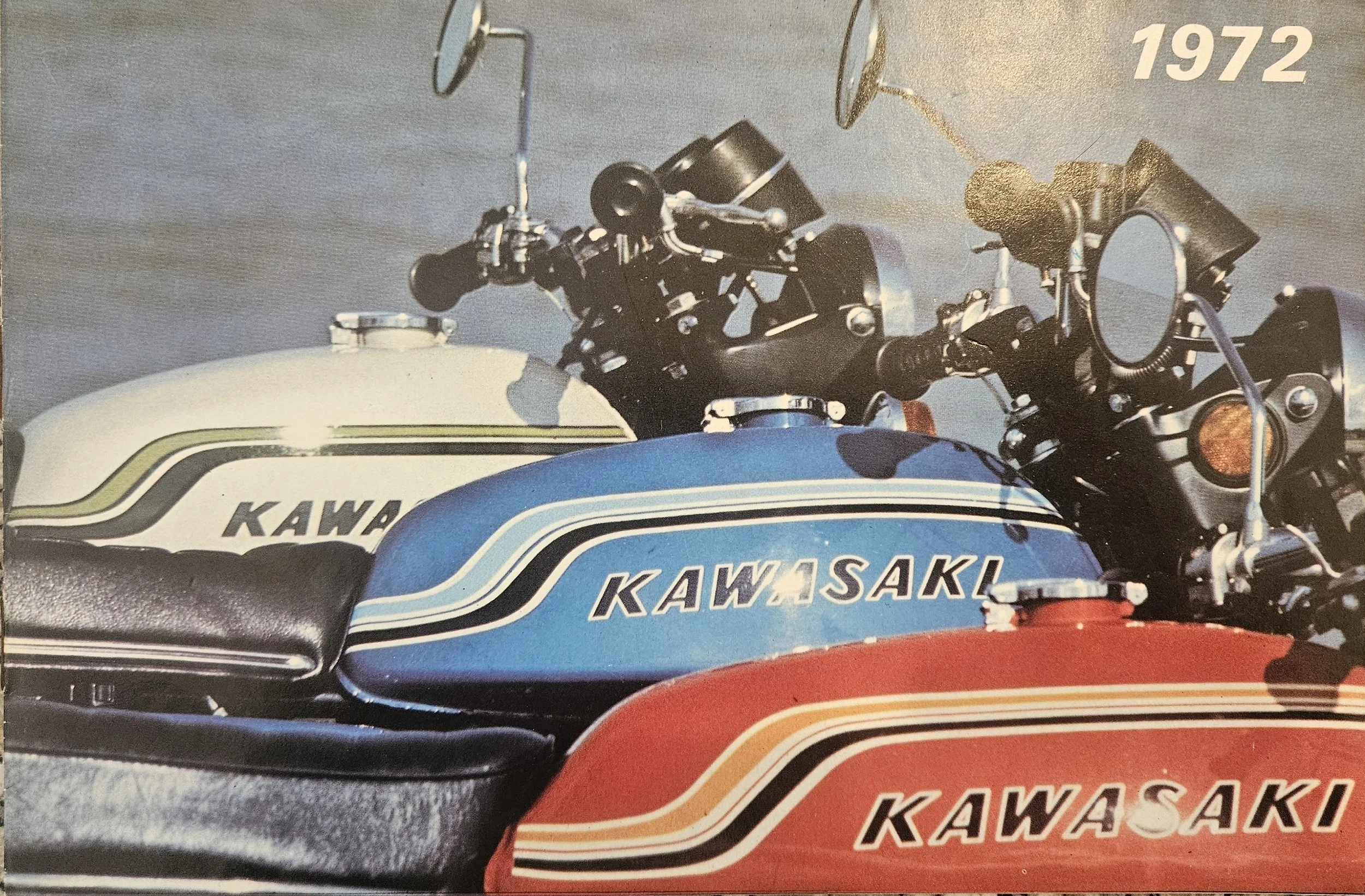 A vintage Kawasaki motorcycle with a blue, white, and red color scheme, featuring the Kawasaki logo on the fuel tank and the year 1972 printed in the top right corner.