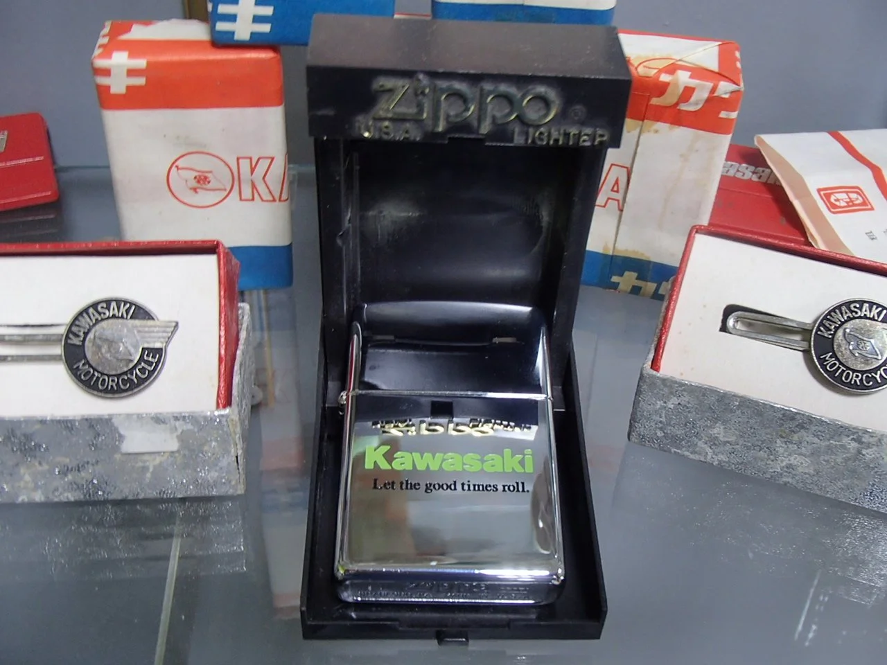 Kawasaki-branded metal lighter inside a black case, surrounded by vintage Kawasaki motorcycle parts and boxes, on a glass surface.