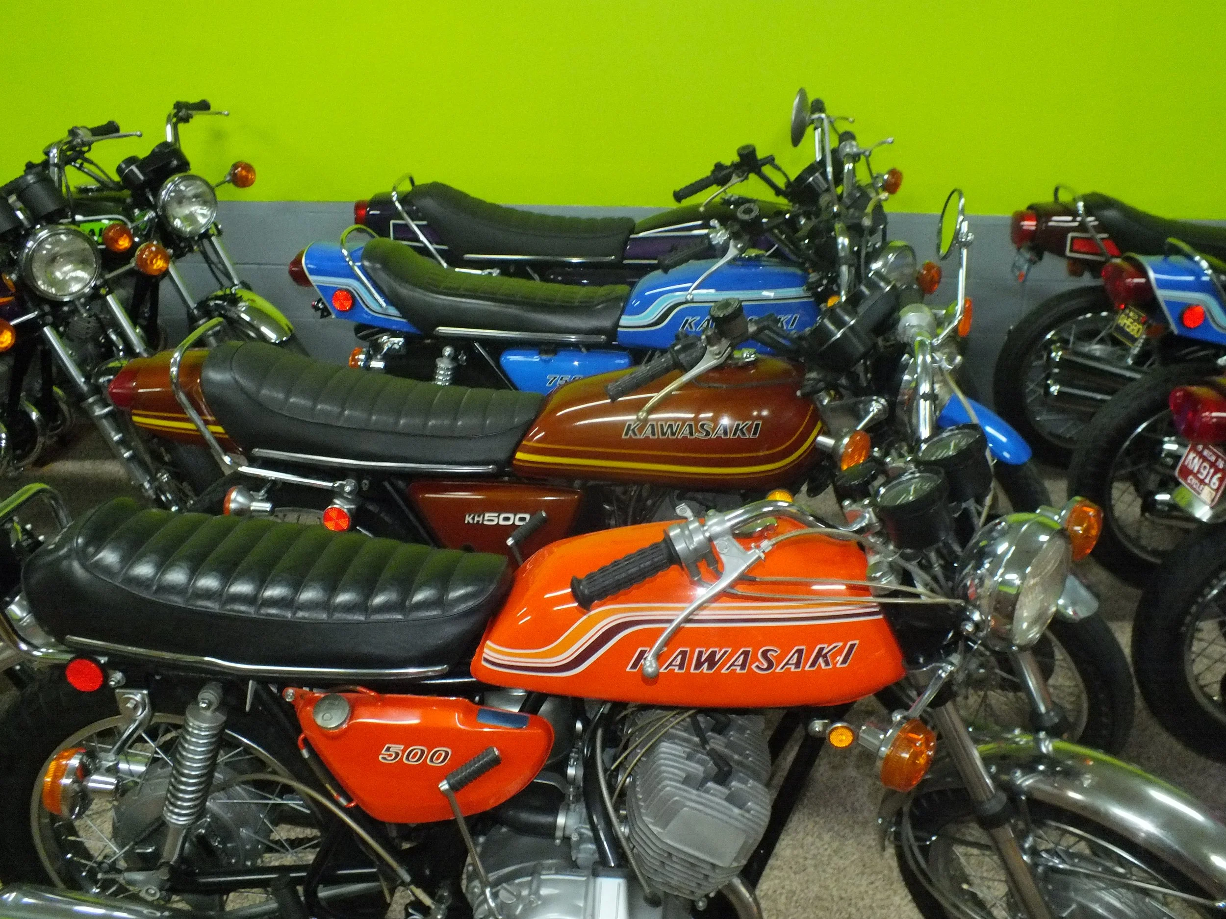 Multiple vintage Kawasaki motorcycles parked indoors against a bright green wall.