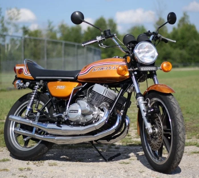 Vintage orange Kawasaki motorcycle parked outdoors on a dirt path with a grassy field and trees in the background.