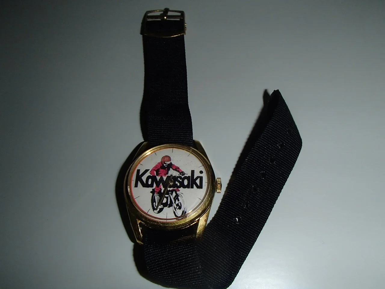 A wristwatch with a black strap and a round face featuring a graphic of a cyclist wearing a red jersey and a helmet, with the word "Kawasaki" in bold black letters behind the cyclist.