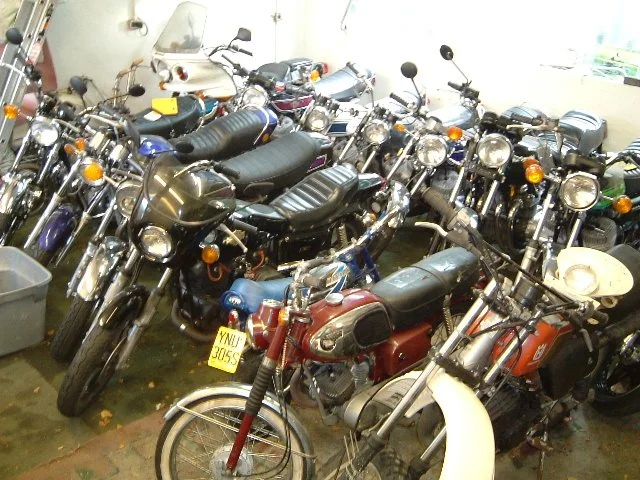 Multiple motorcycles and scooters parked indoors, some with helmets, in a cluttered storage or garage space.