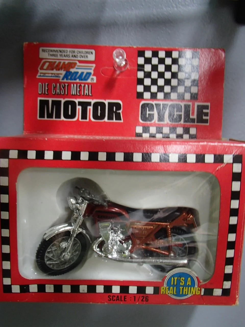 A die-cast metal motorcycle toy in a red box with checkered patterns. The box has labels for 'Motor Cycle' and 'It's a real thing', with a scale of 1/26. The motorcycle is metallic with a chrome finish and black tires.
