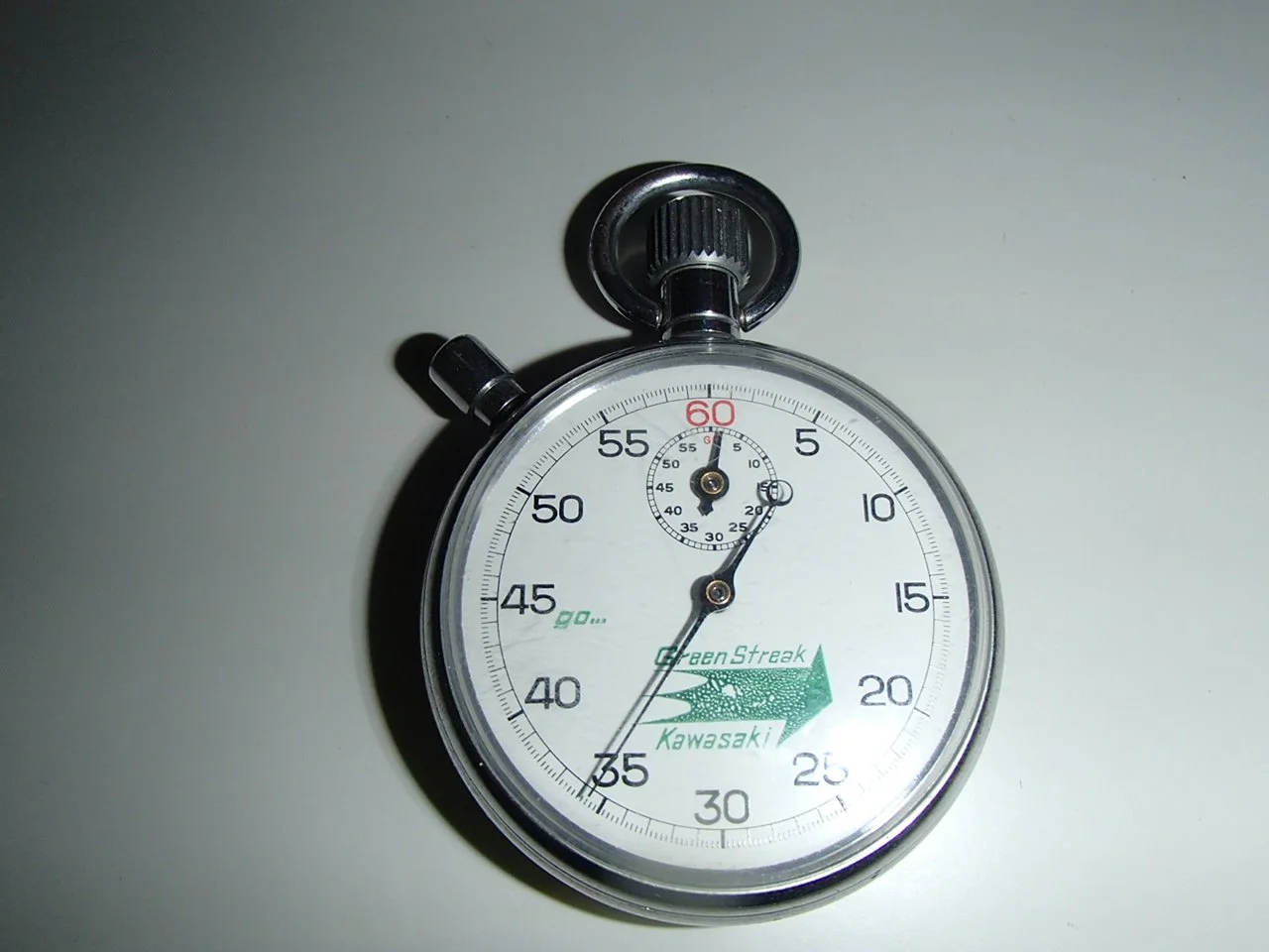 A silver stopwatch with a white face, black numbers, and green text and arrow, showing approximately 37 seconds, with a small top dial indicating the seconds, and branding for GreenStreak and Kawasaki.