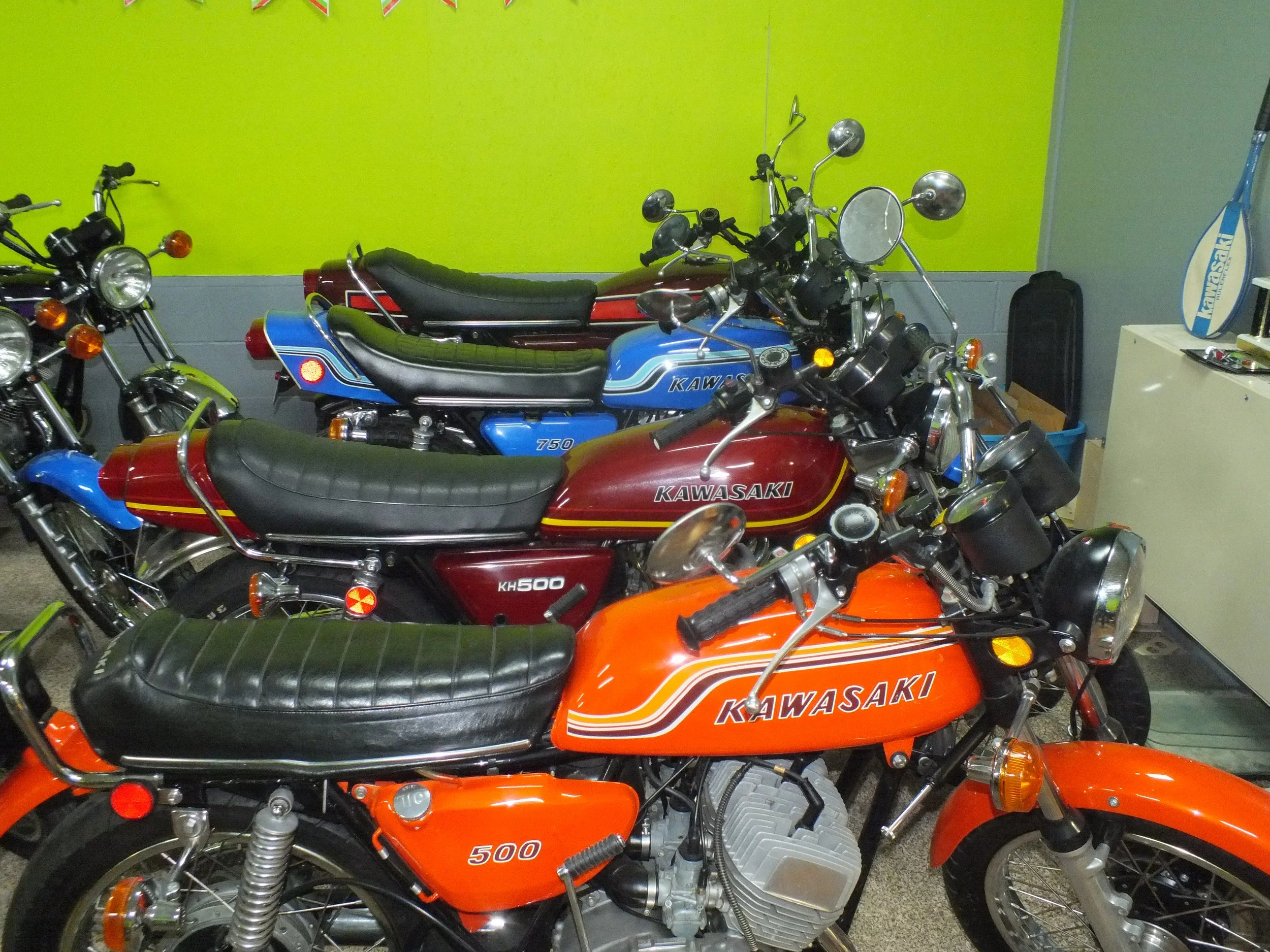 A collection of vintage Kawasaki motorcycles parked indoors with a green wall and bunting in the background.