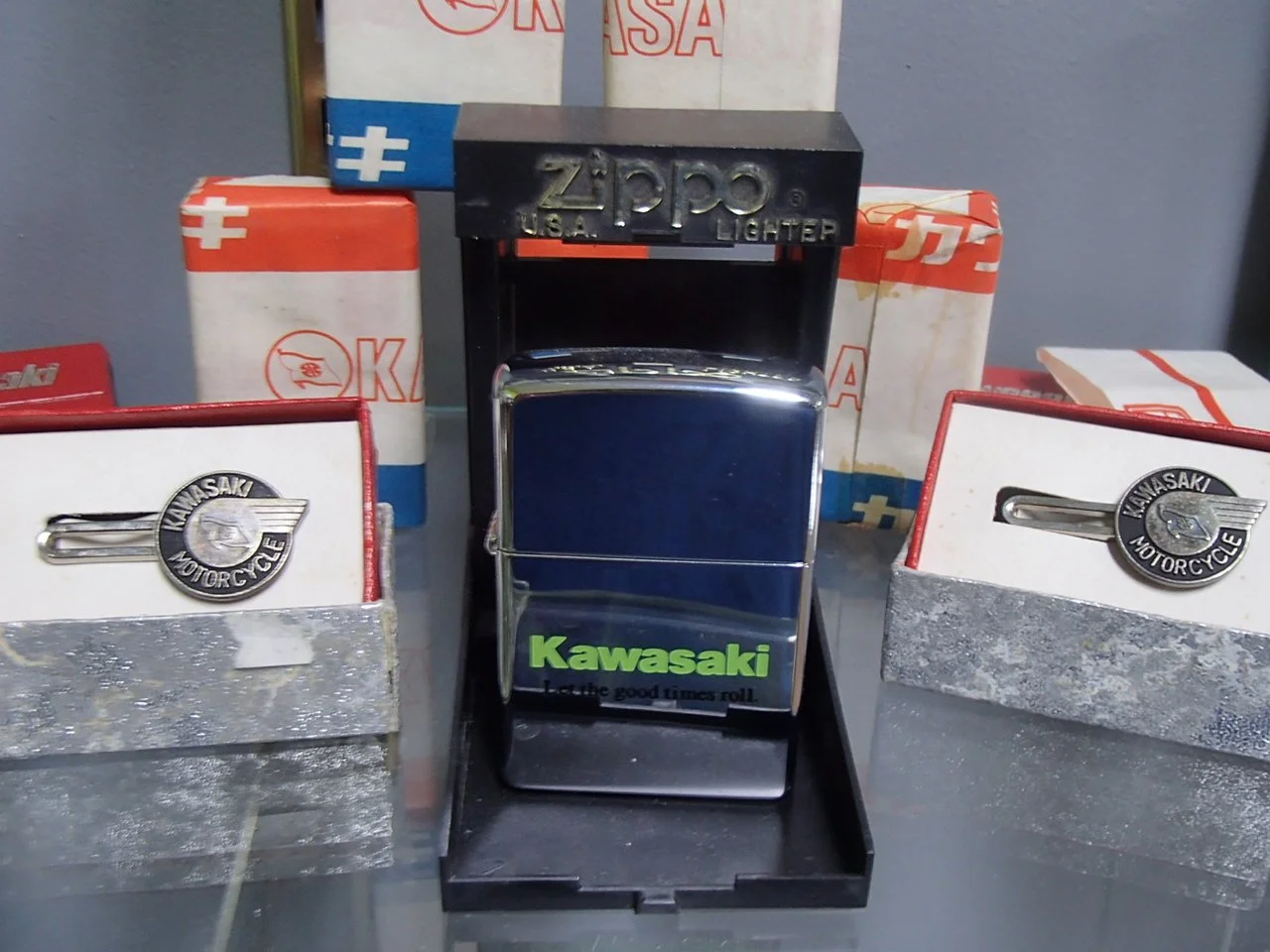 A vintage Zippo lighter display with Kawasaki motorcycle insignia, surrounded by vintage Kawasaki cigarette lighters in original packaging.