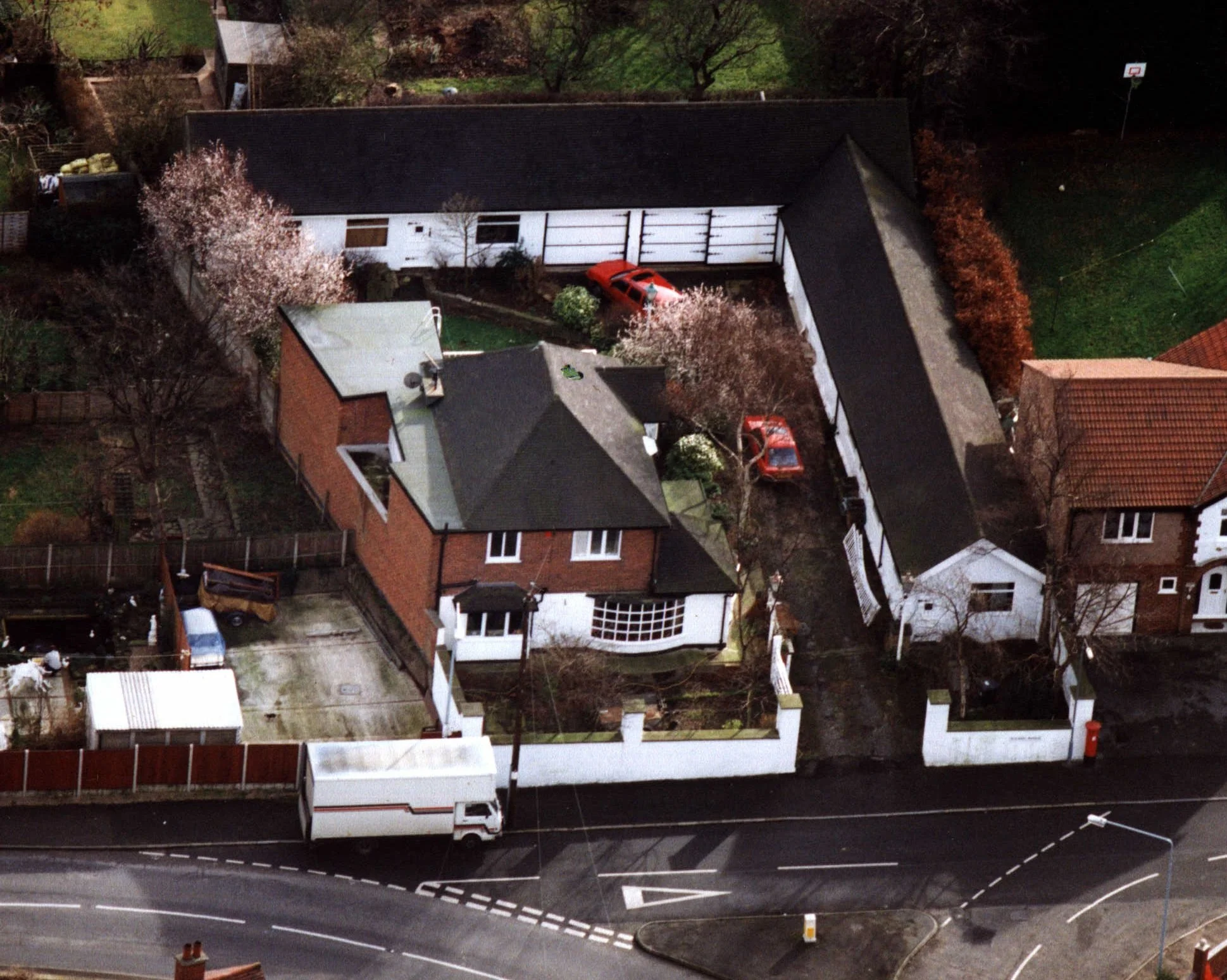 An aerial view of a residential neighborhood at night, showing some houses, cars parked in driveways and streets, trees, and a basketball hoop. A white truck is parked on the street in the foreground.