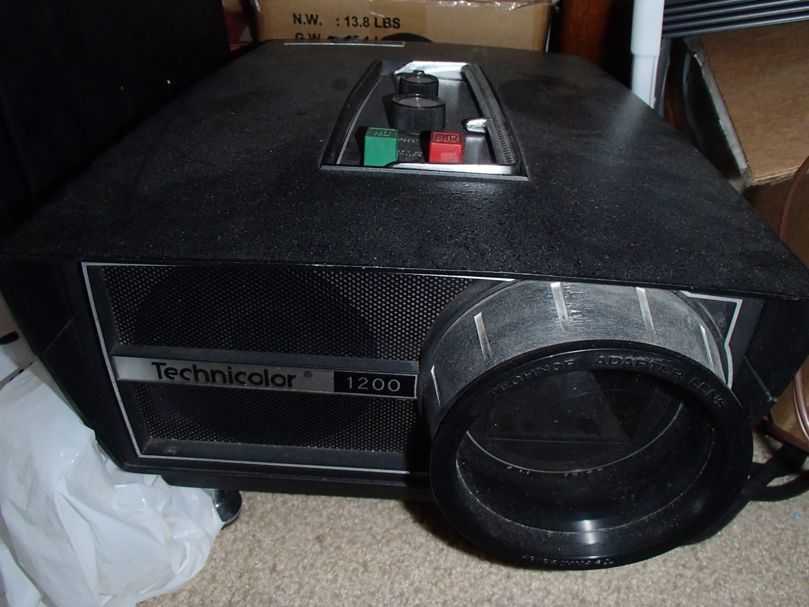 A vintage Technicolor 1200 slide projector with a black textured surface, silver accents, and a large lens. There are three controls on top and it is placed on a carpeted floor next to a white plastic bag and some cardboard boxes.
