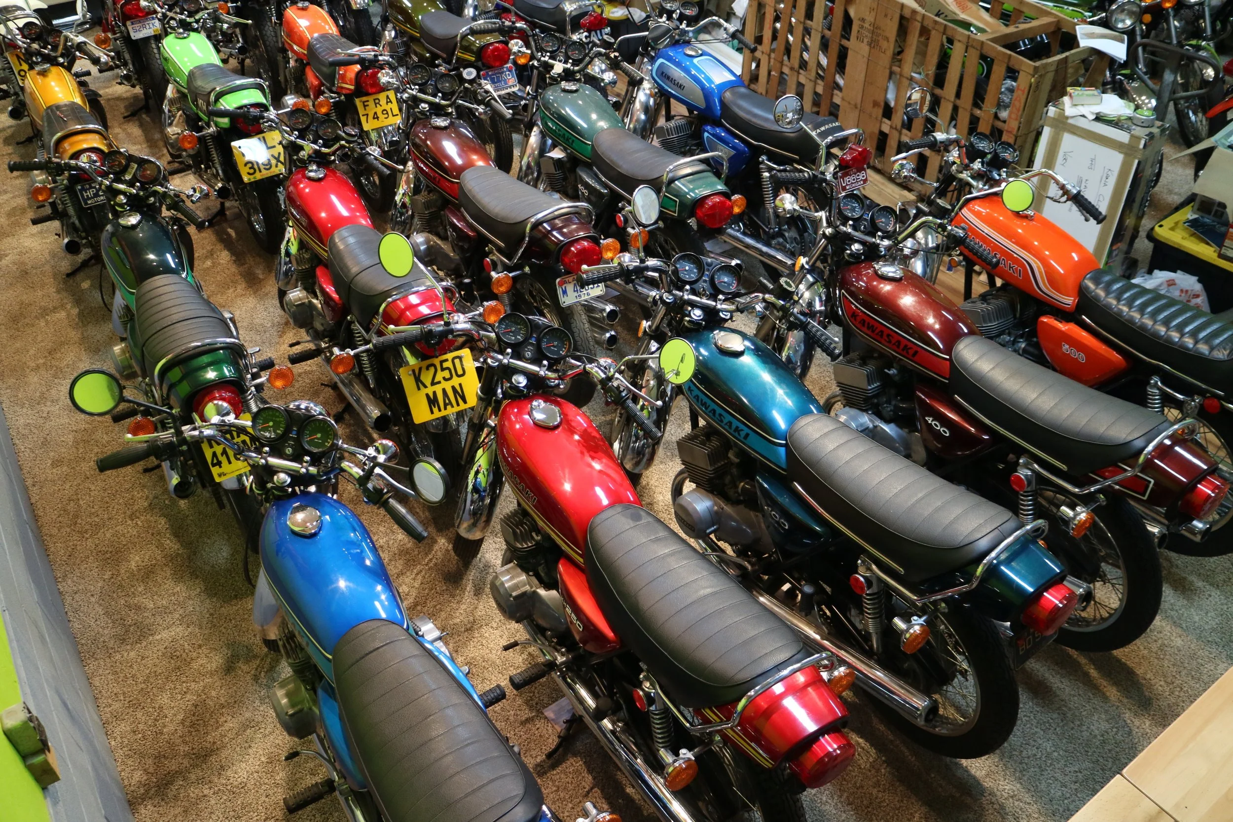 Multiple vintage Kawasaki motorcycles parked indoors, featuring red, blue, green, and orange colors with speedometers, mirrors, and license plates visible.