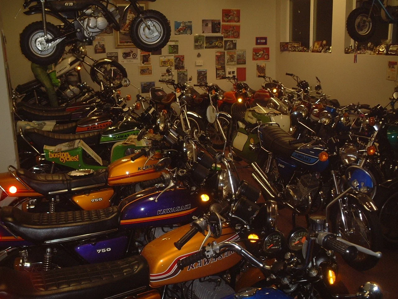 Room filled with vintage Kawasaki motorcycles, some hanging from the ceiling, and posters on the wall.