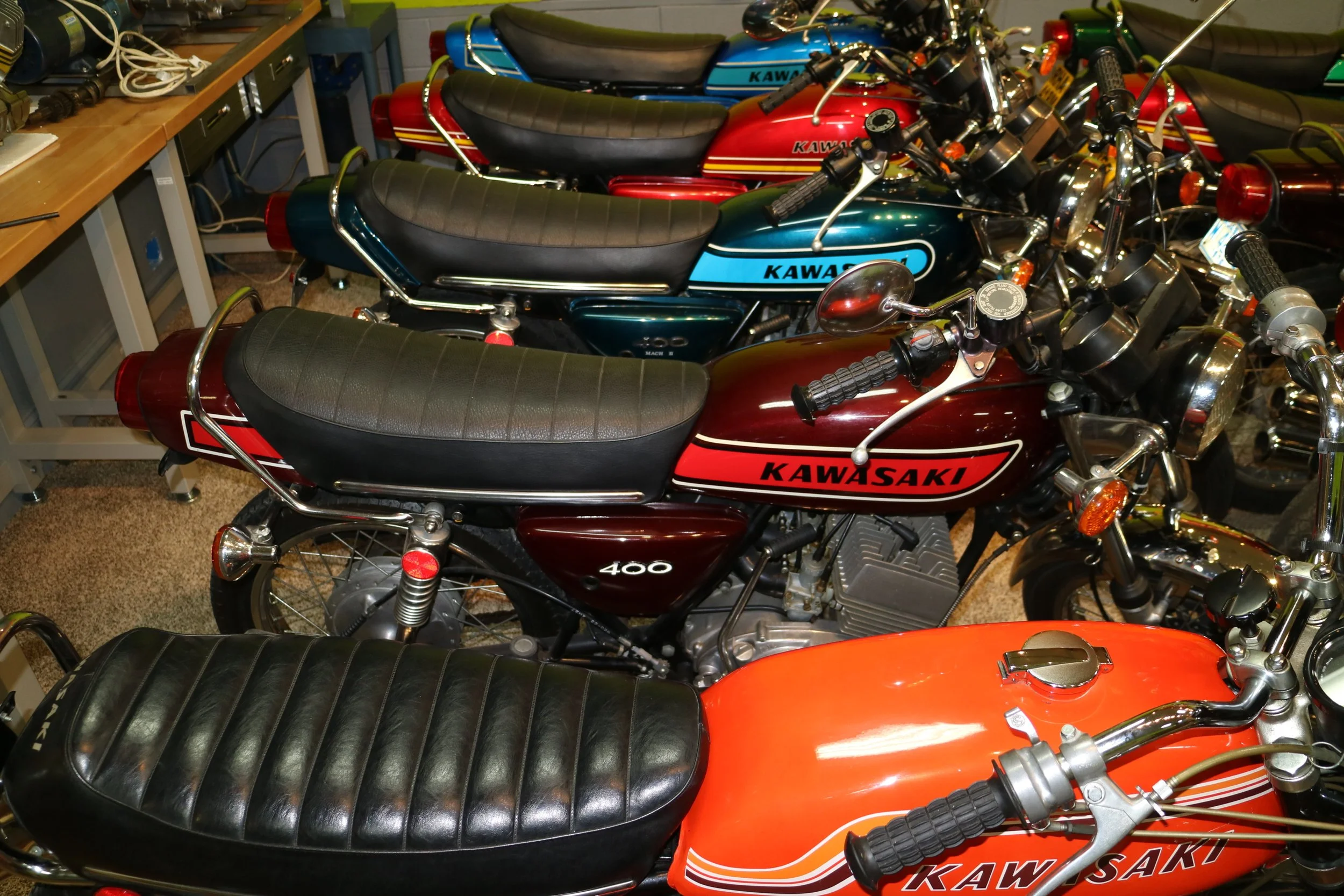 Multiple vintage Kawasaki motorcycles parked indoors, with black leather seats and chrome details, in a workshop or garage setting.
