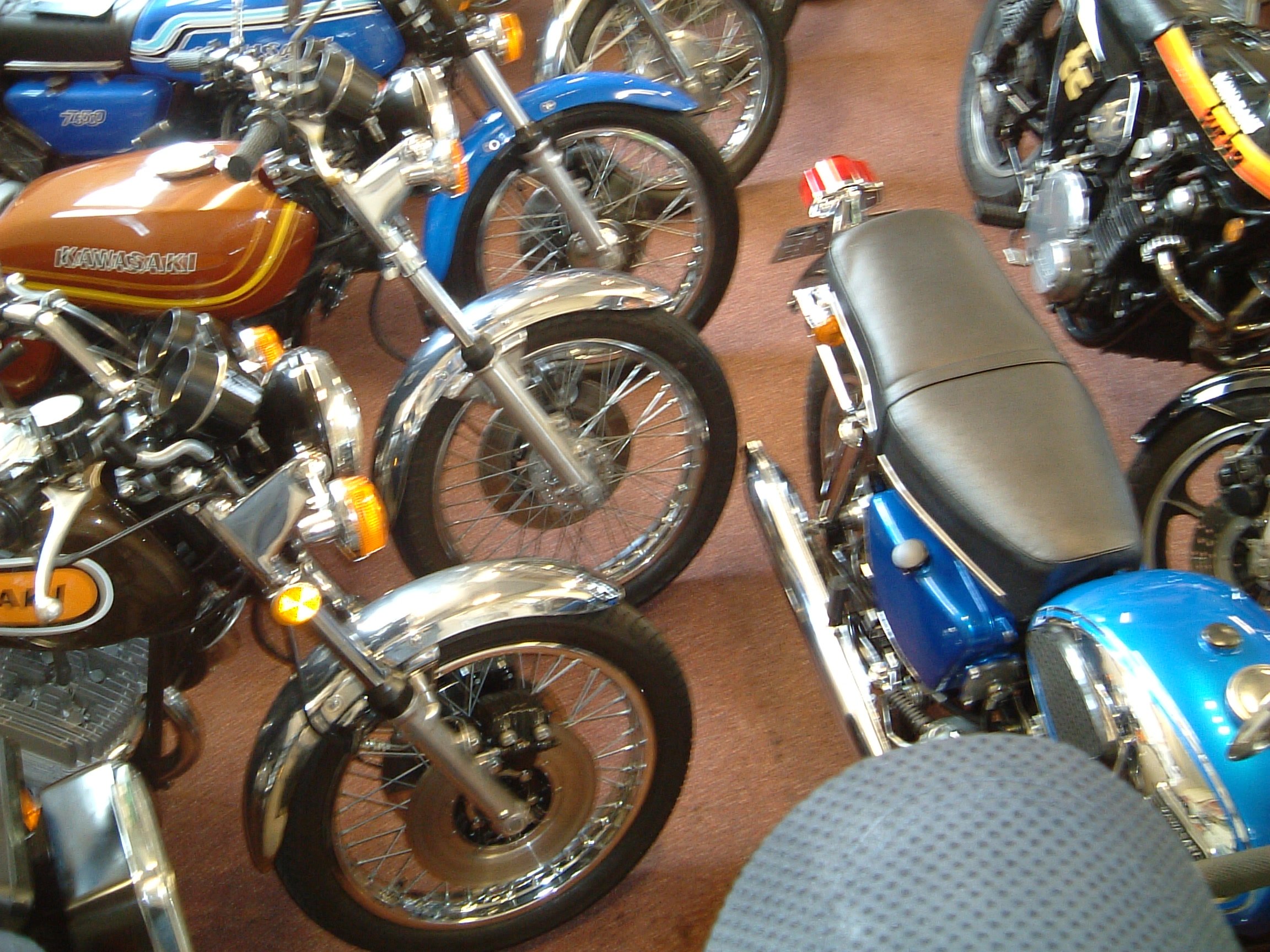 Multiple motorcycles on display, including a blue Kawasaki, a brown Kawasaki, and a blue bike. The motorcycles are parked close together on a brown carpeted floor.