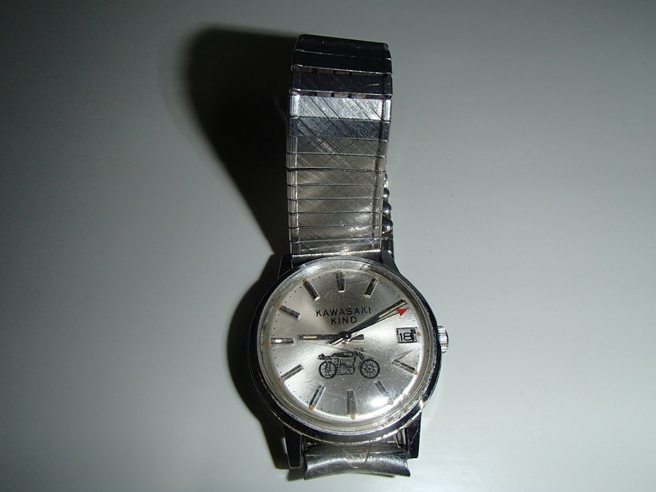 A vintage Kawasaki Kind wristwatch with a silver band, white dial, date window, and a small motorcycle illustration on the face, placed on a plain surface.