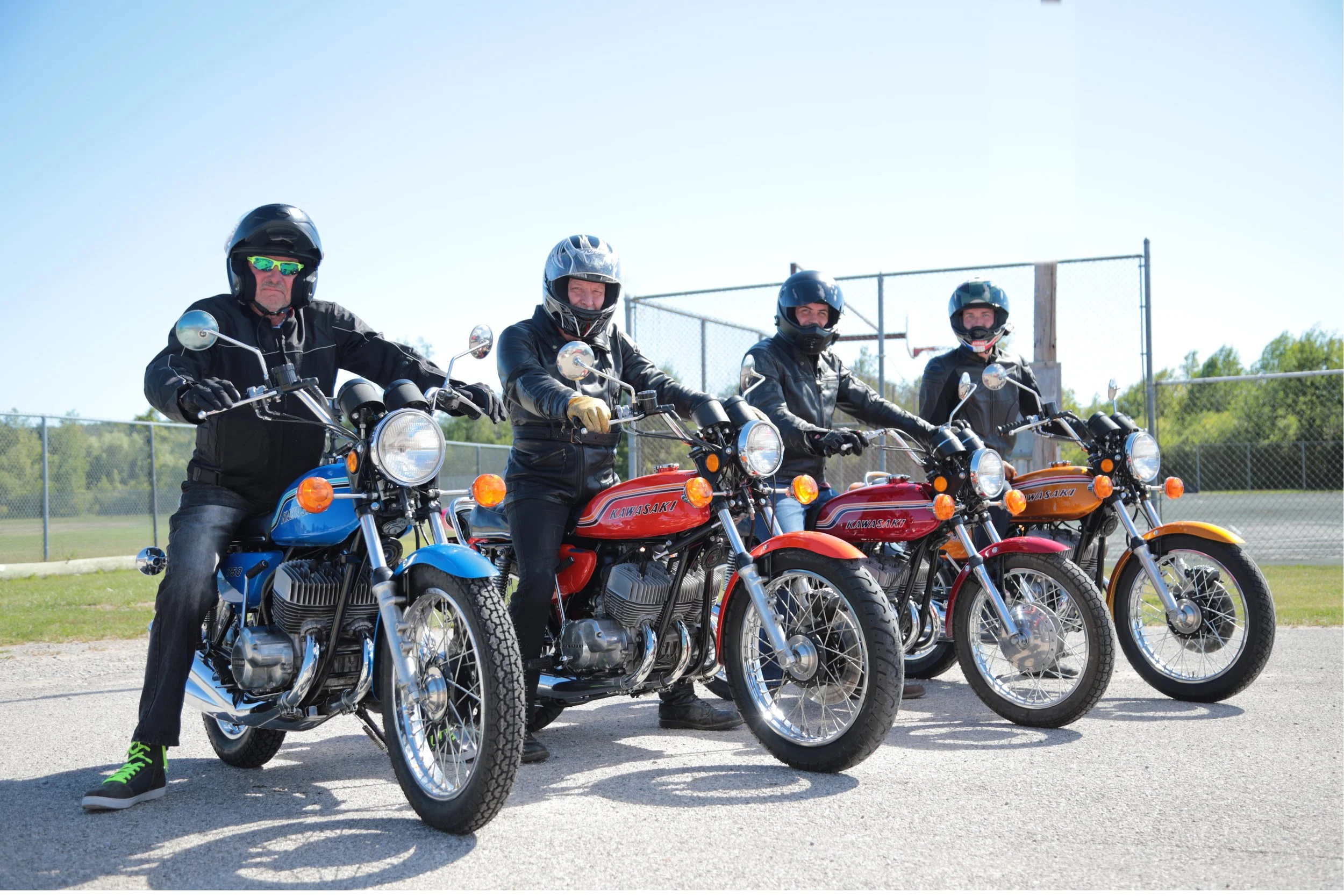 Four people on vintage Kawasaki motorcycles lined up outdoors on a sunny day, all wearing helmets and jackets, with a chain-link fence and trees in the background.