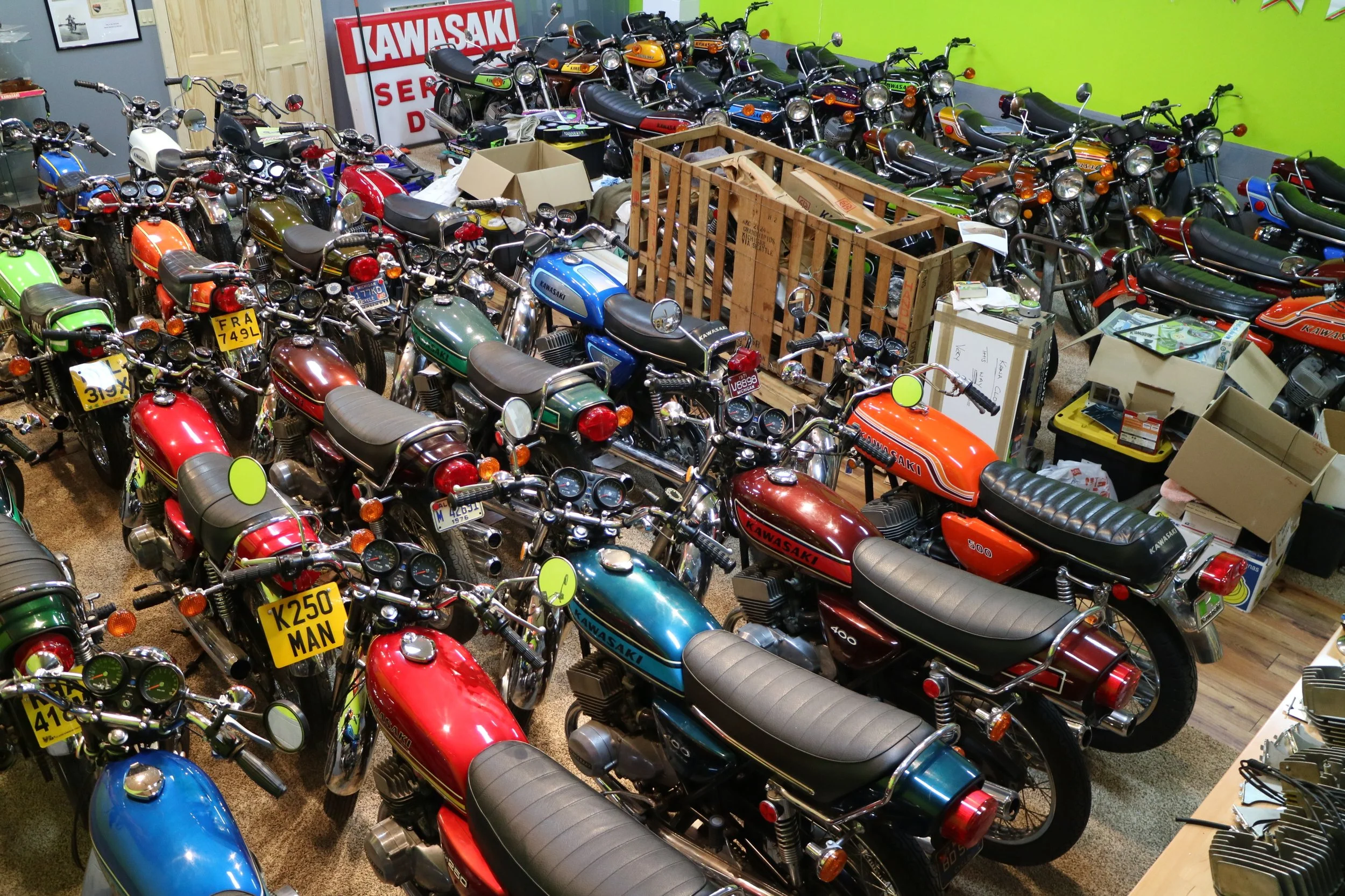 A room filled with numerous vintage Kawasaki motorcycles parked closely together, with some boxes and items in the middle and a bright green wall on one side.
