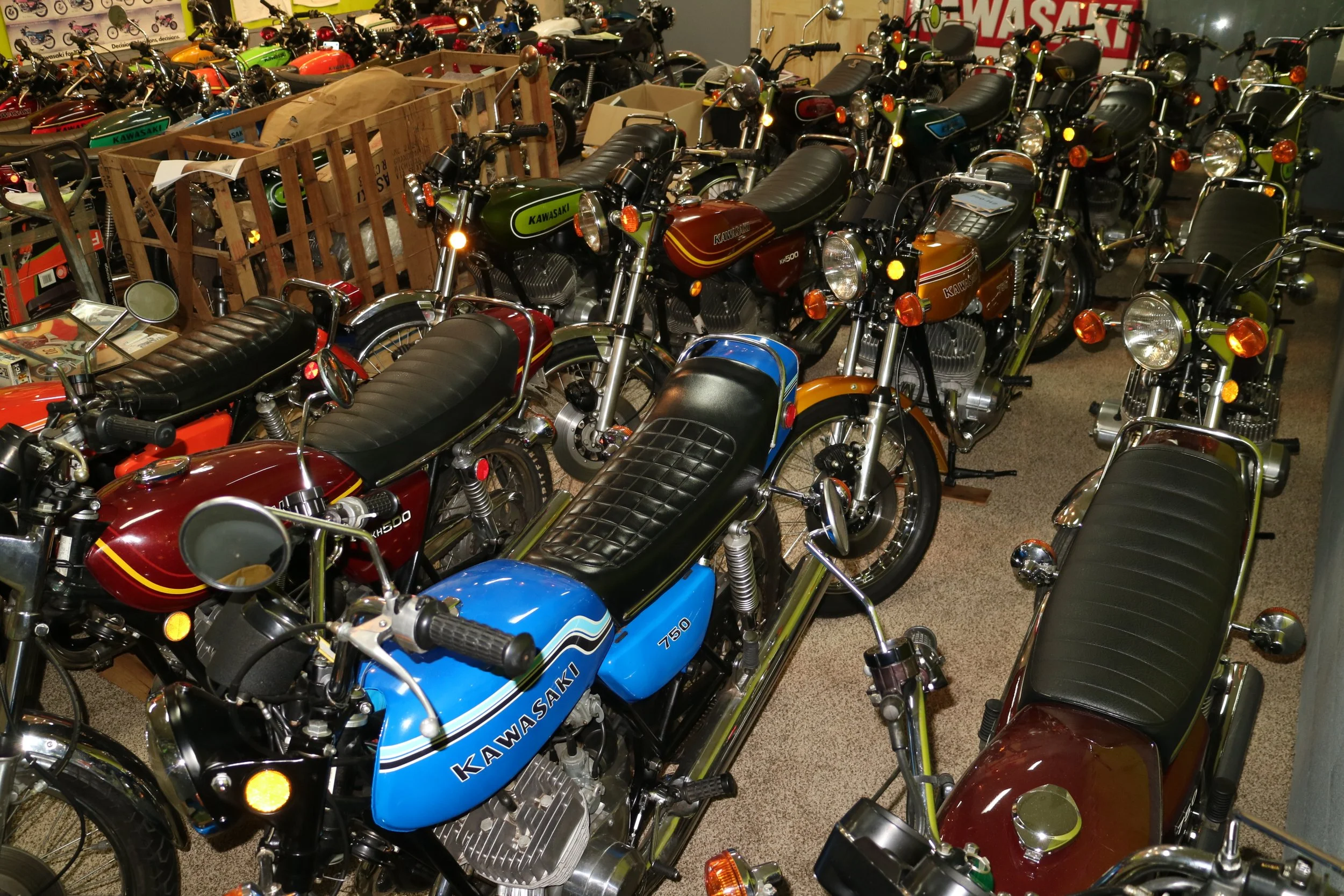 A collection of vintage Kawasaki motorcycles and minibikes displayed in an indoor showroom or storage space, with various colors including green, blue, orange, and brown.