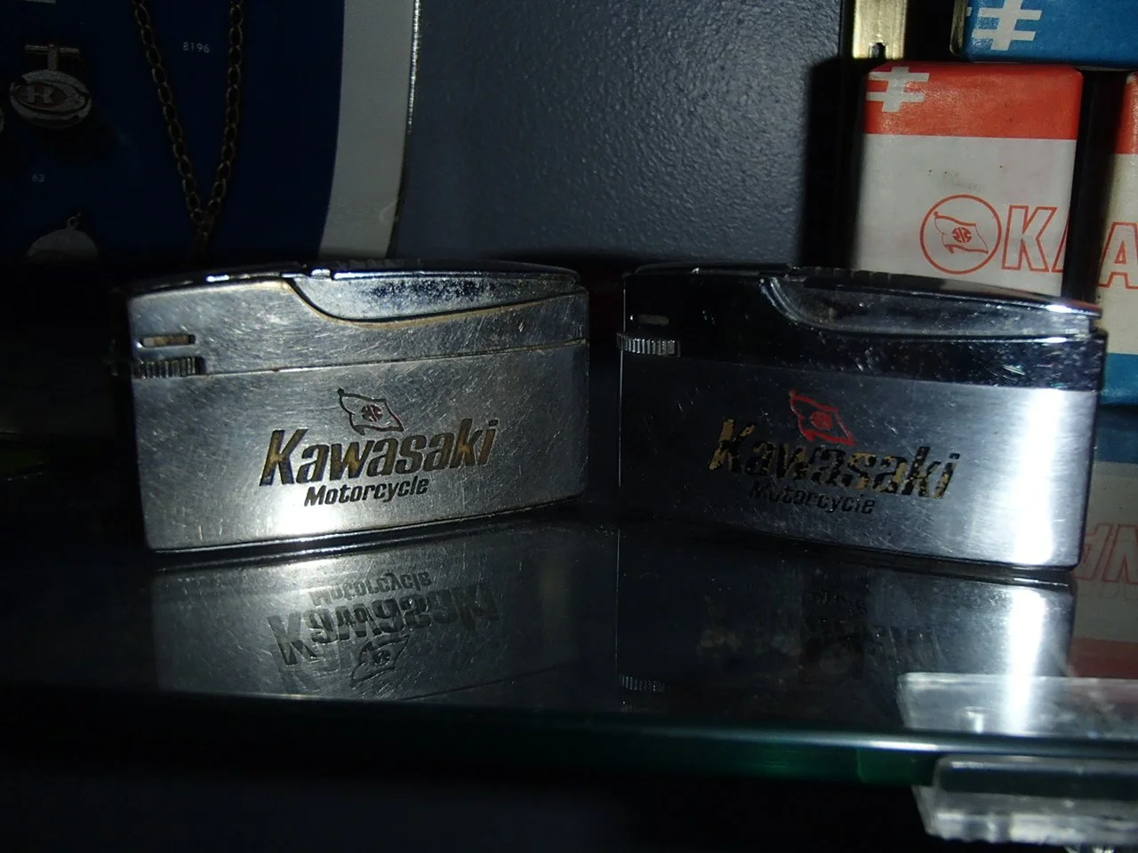 Two vintage metal Kawasaki motorcycle tins on a glass surface, with boxes and other items in the background.