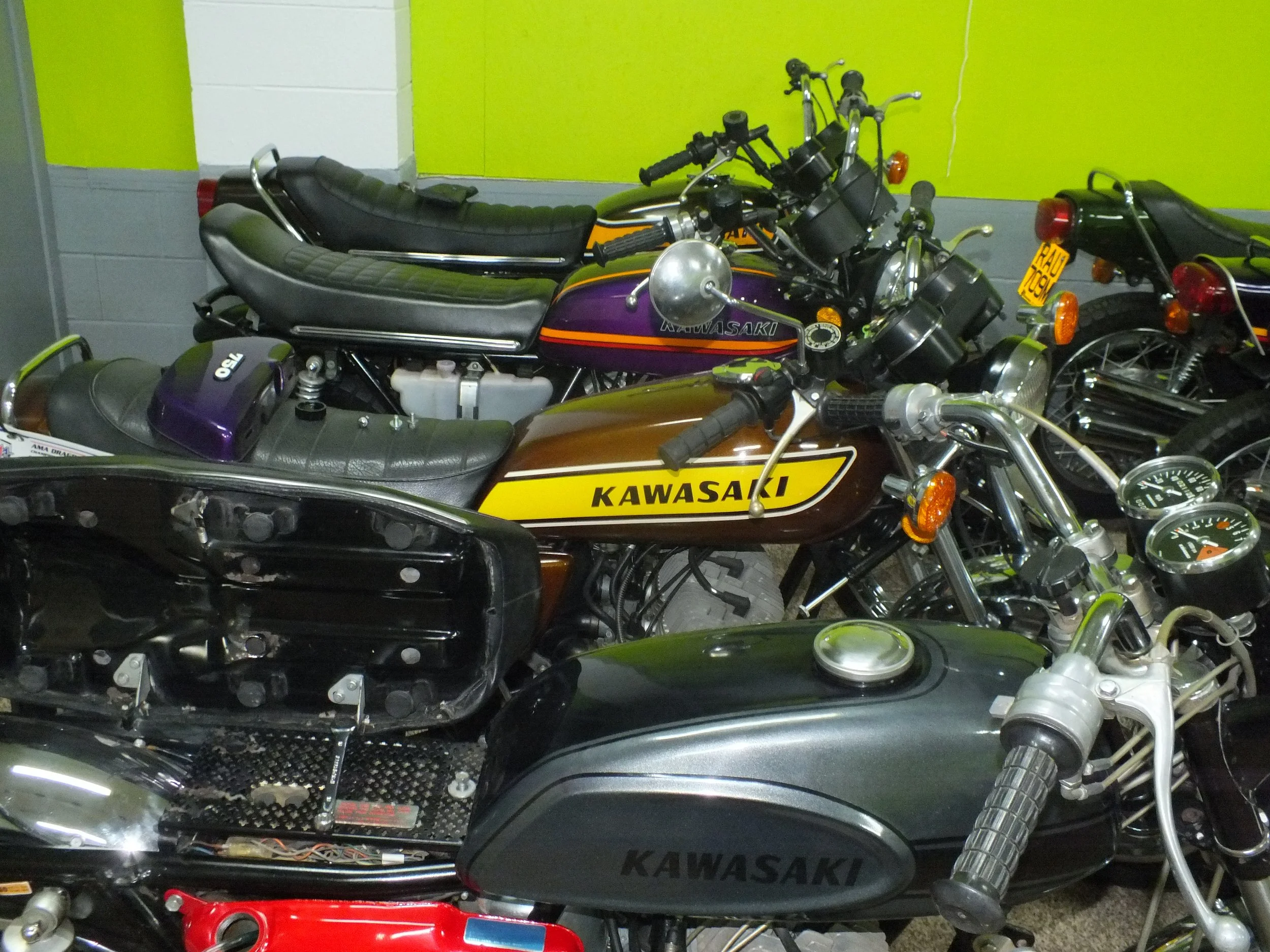 Multiple motorcycles parked indoors, some with Kawasaki branding, with one bike's seat and body parts partially disassembled.