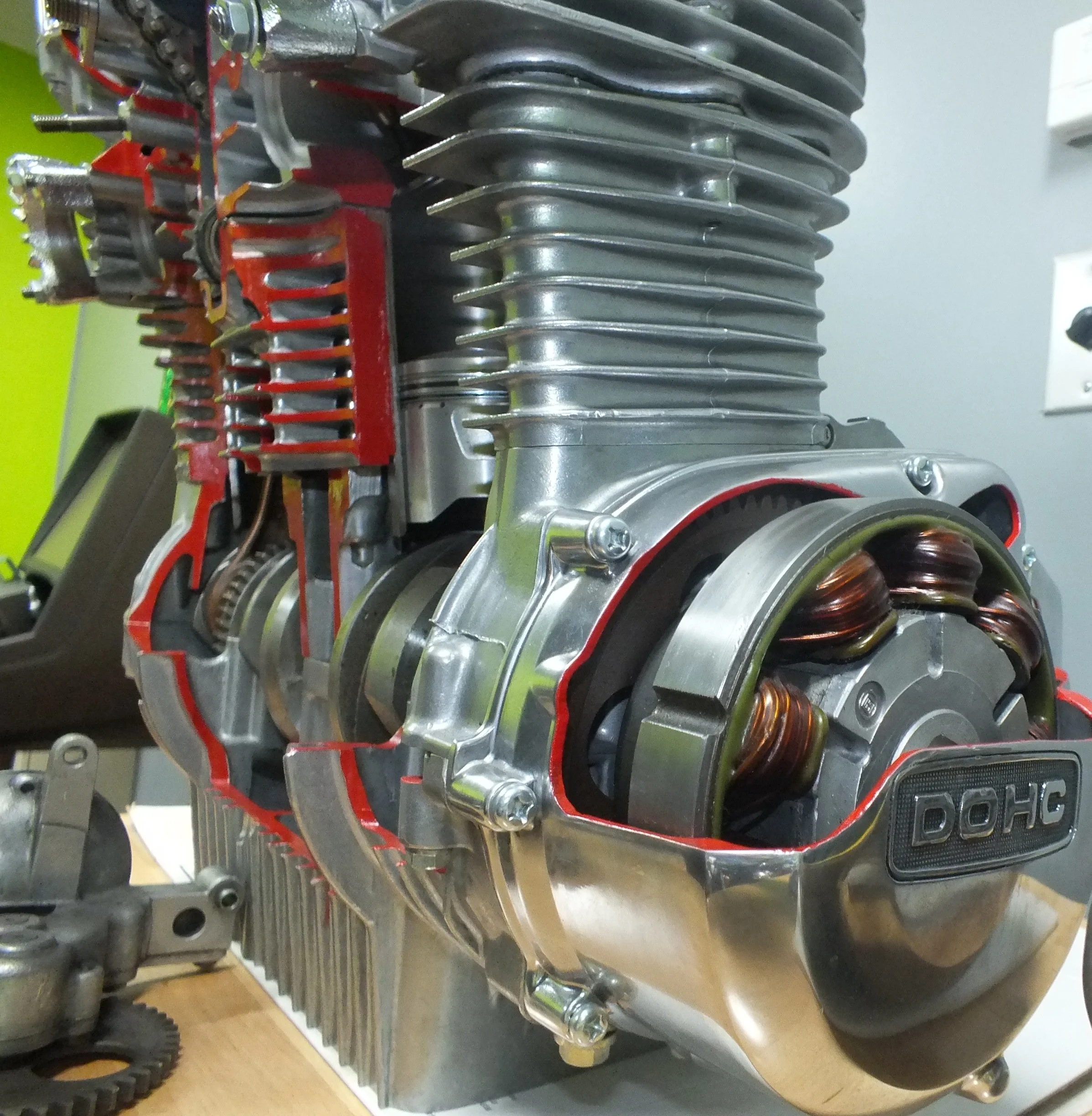 Close-up of a vintage single-cylinder engine with cooling fins, part of a motorcycle or similar vehicle, showing the cylinder head, spark plug, and coil spring on a light-colored surface with a green wall in the background.