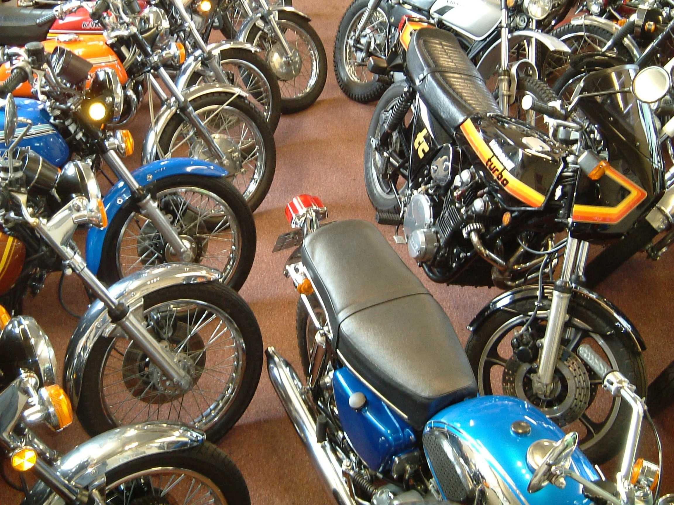 A collection of vintage motorcycles and mopeds in various colors including blue, orange, black, and silver, displayed indoors on a brown carpeted floor.