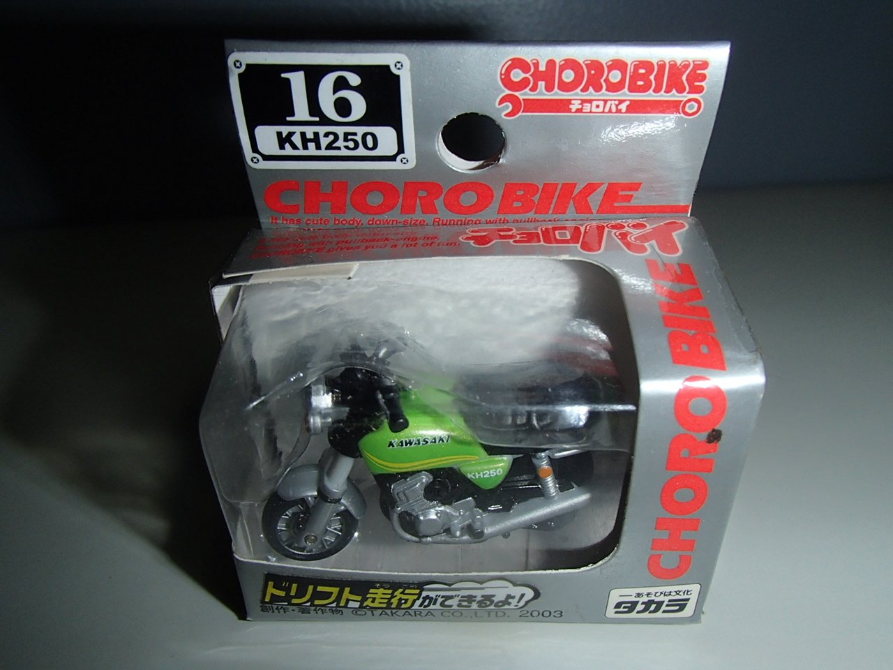 A small green Kawasaki KH250 motorcycle toy inside a clear plastic display box with red and white text on the packaging.
