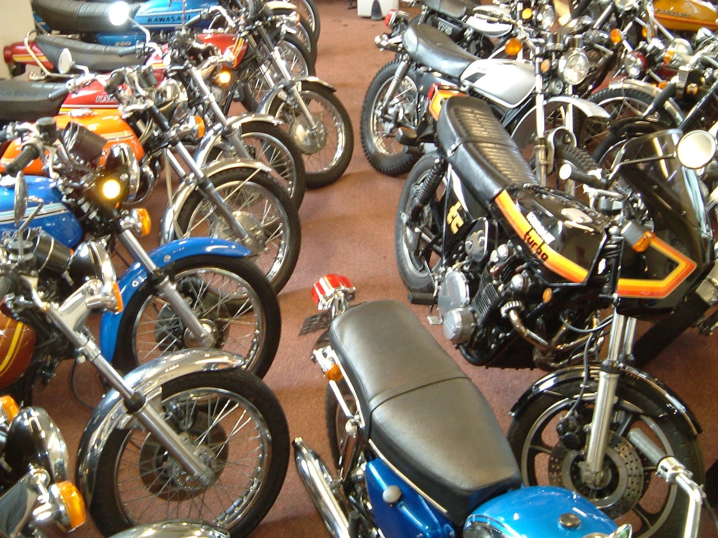 Multiple vintage motorcycles and scooters parked indoors, with some having bright orange, blue, and silver colors.