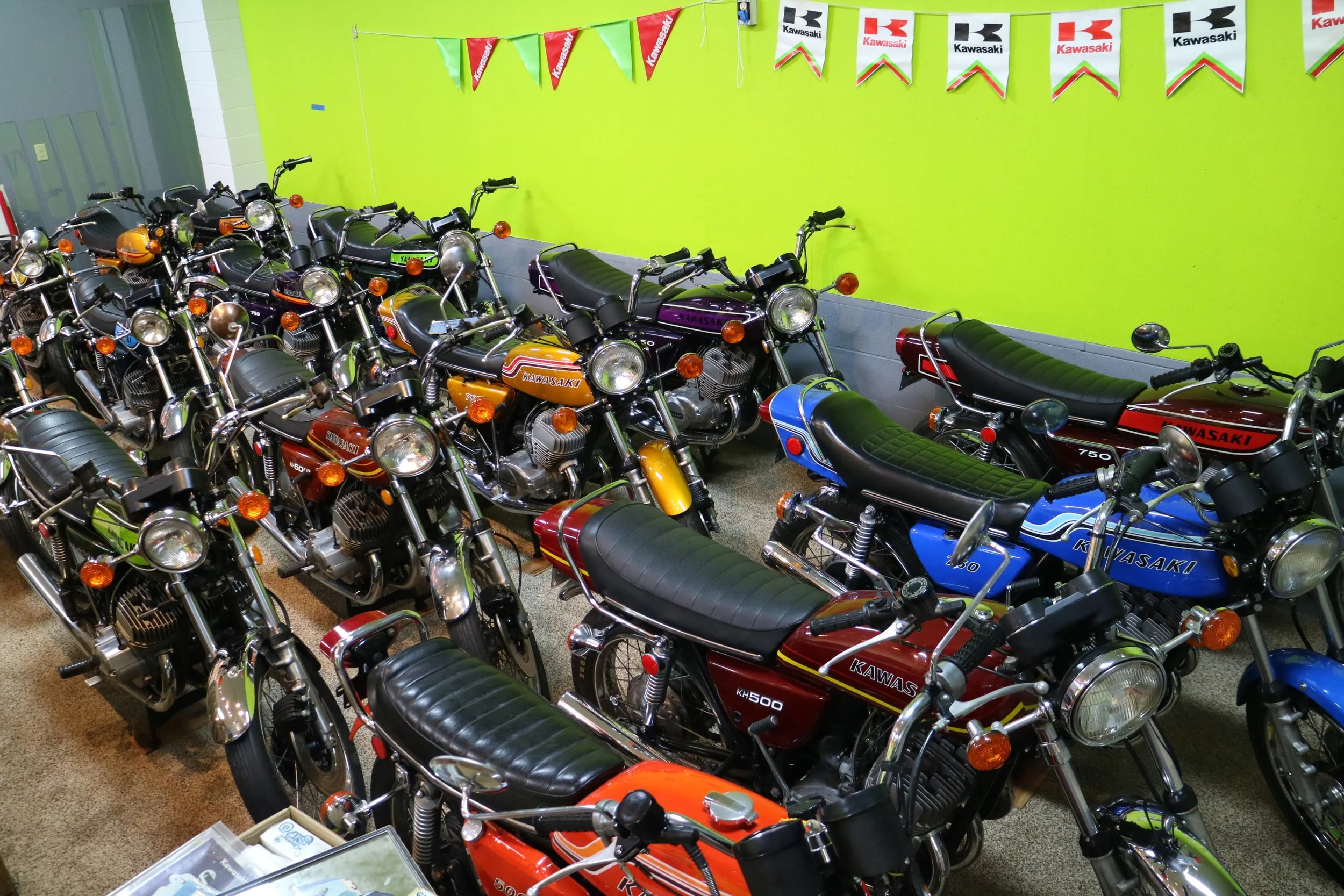 A showroom filled with vintage Kawasaki motorcycles arranged in rows against bright yellow and green walls, with Kawasaki banners hanging overhead.