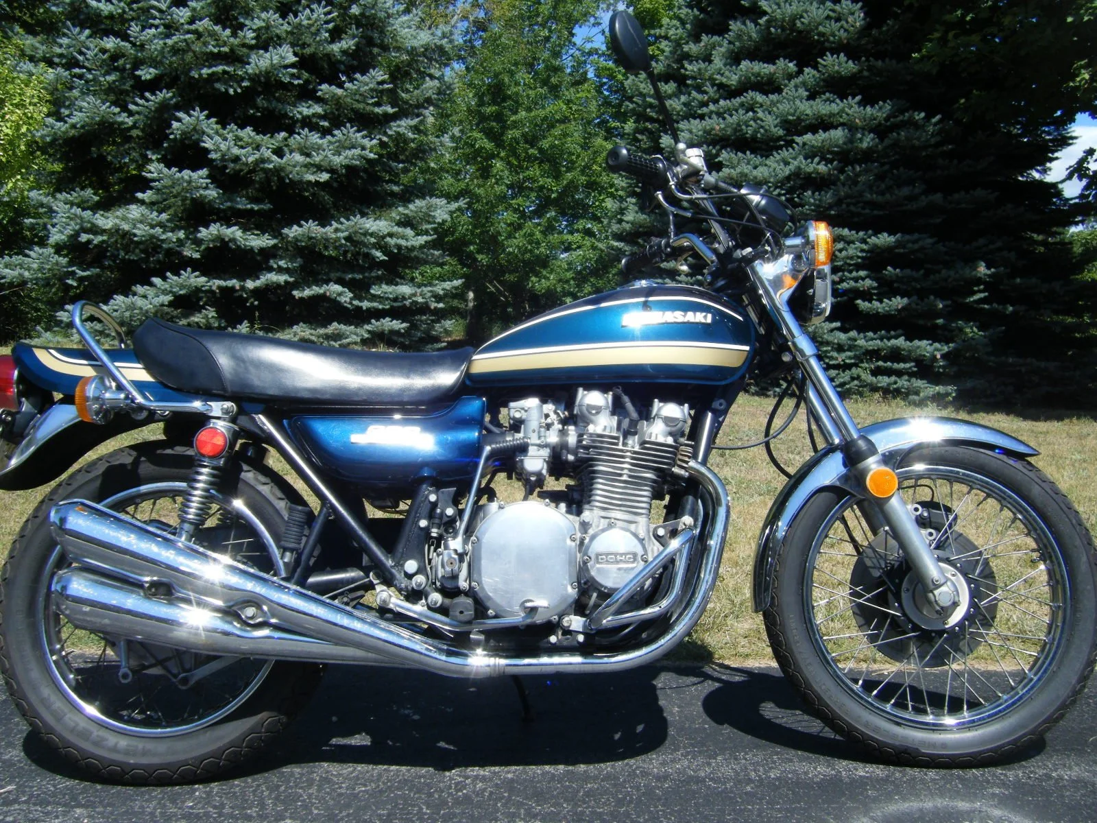 A vintage blue Kawasaki motorcycle with chrome accents, parked outdoors on a paved surface, with green trees and grass in the background.