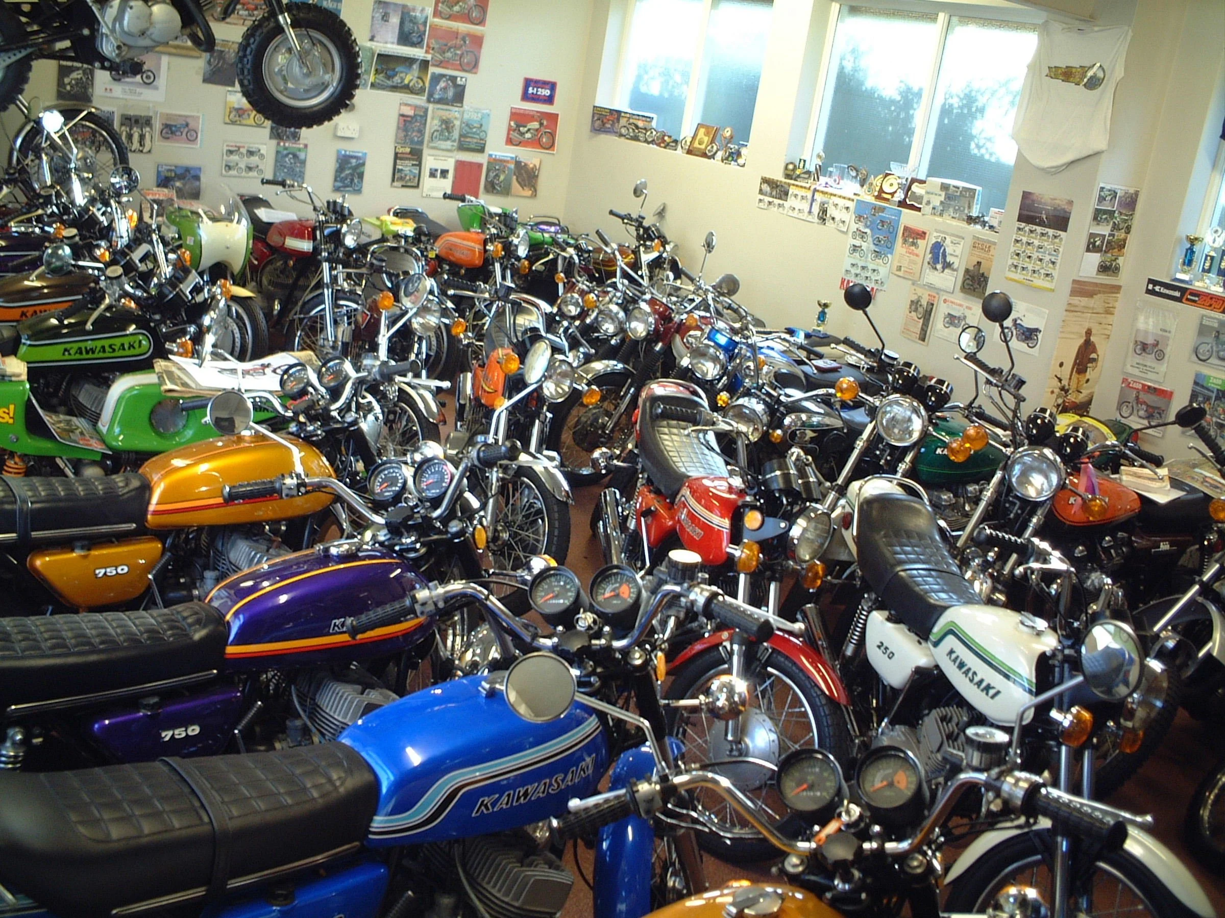 A room filled with multiple vintage Kawasaki motorcycles parked closely together. The room has posters and model pictures of motorcycles on the walls, with natural light coming from windows.