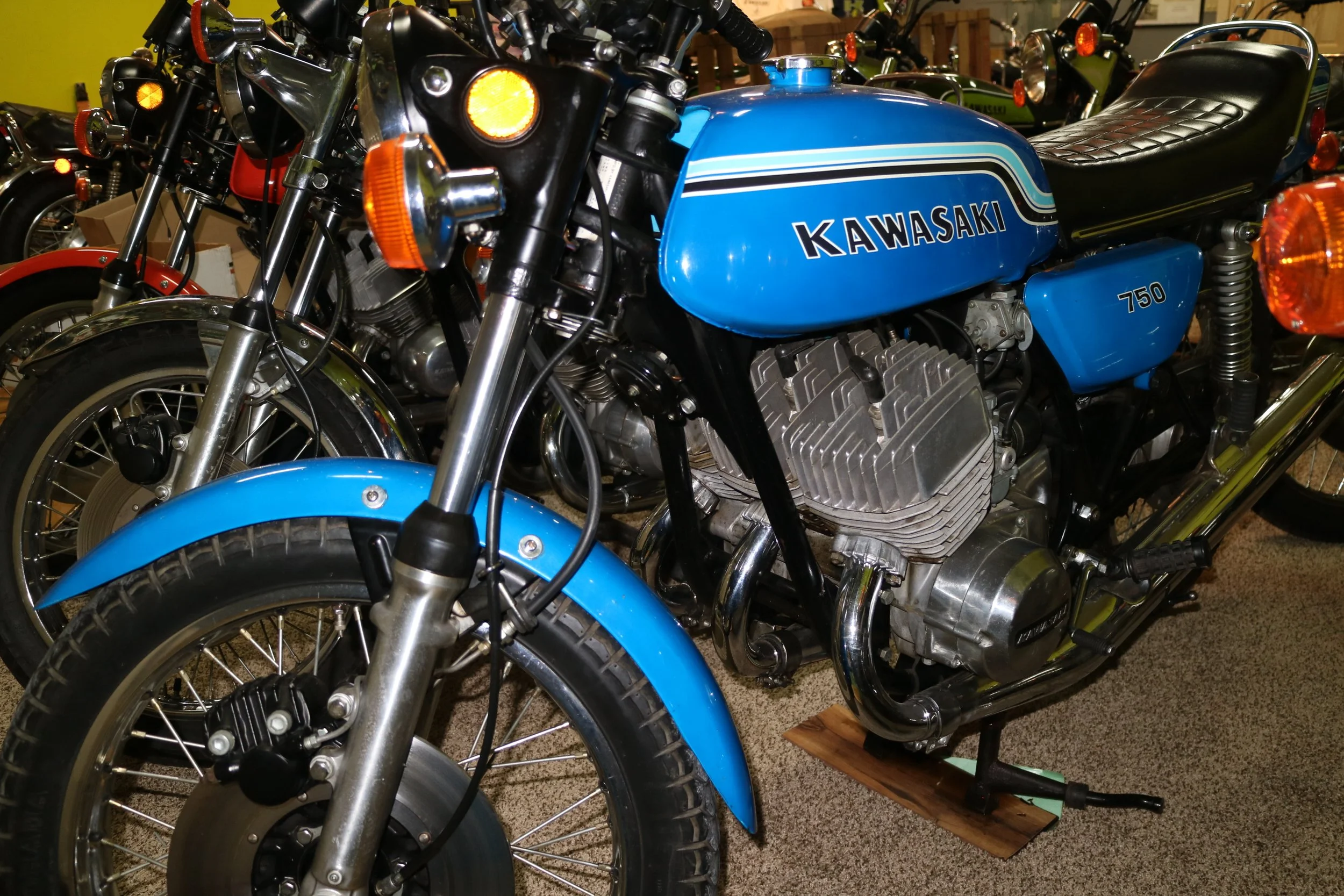 A blue Kawasaki motorcycle with a black leather seat and chrome details is displayed among other motorcycles in a showroom or museum. The motorcycle has a classic design, with a visible engine and a front wheel with spokes.