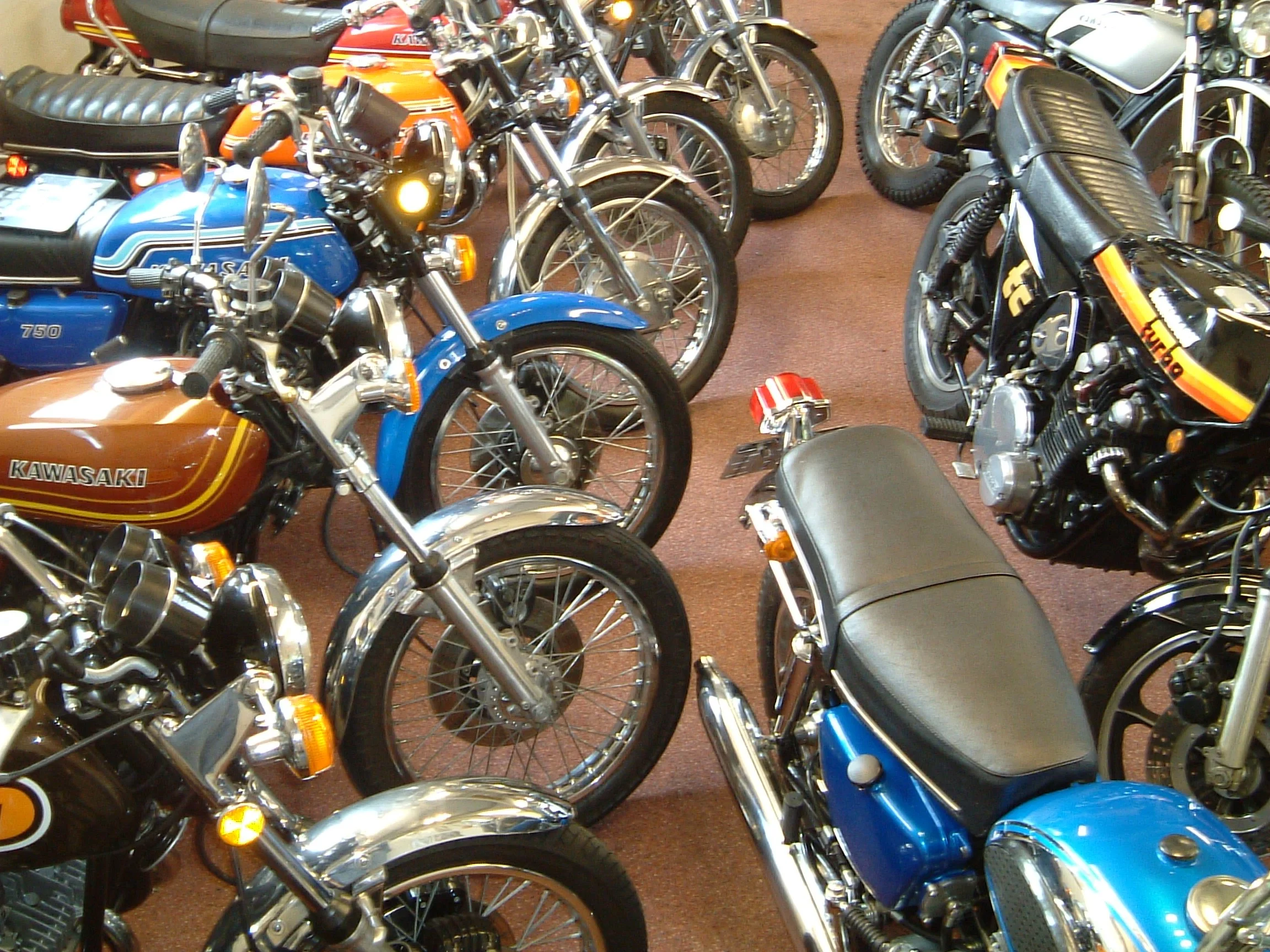 A collection of vintage motorcycles indoors, featuring Kawasaki, Honda, and Yamaha models with various colors and styles.