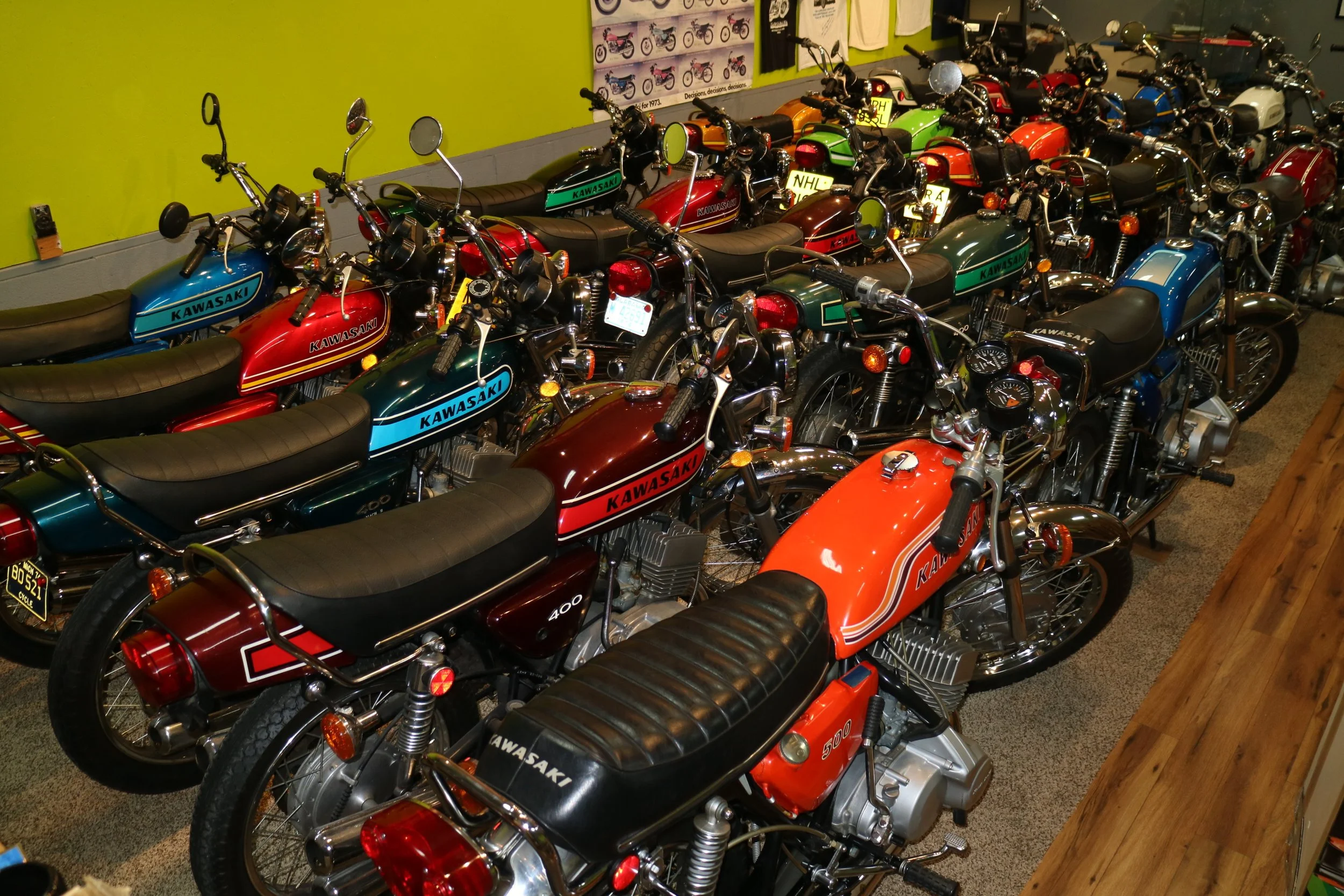 Multiple vintage Kawasaki motorcycles lined up inside a showroom with colorful walls and promotional posters in the background.