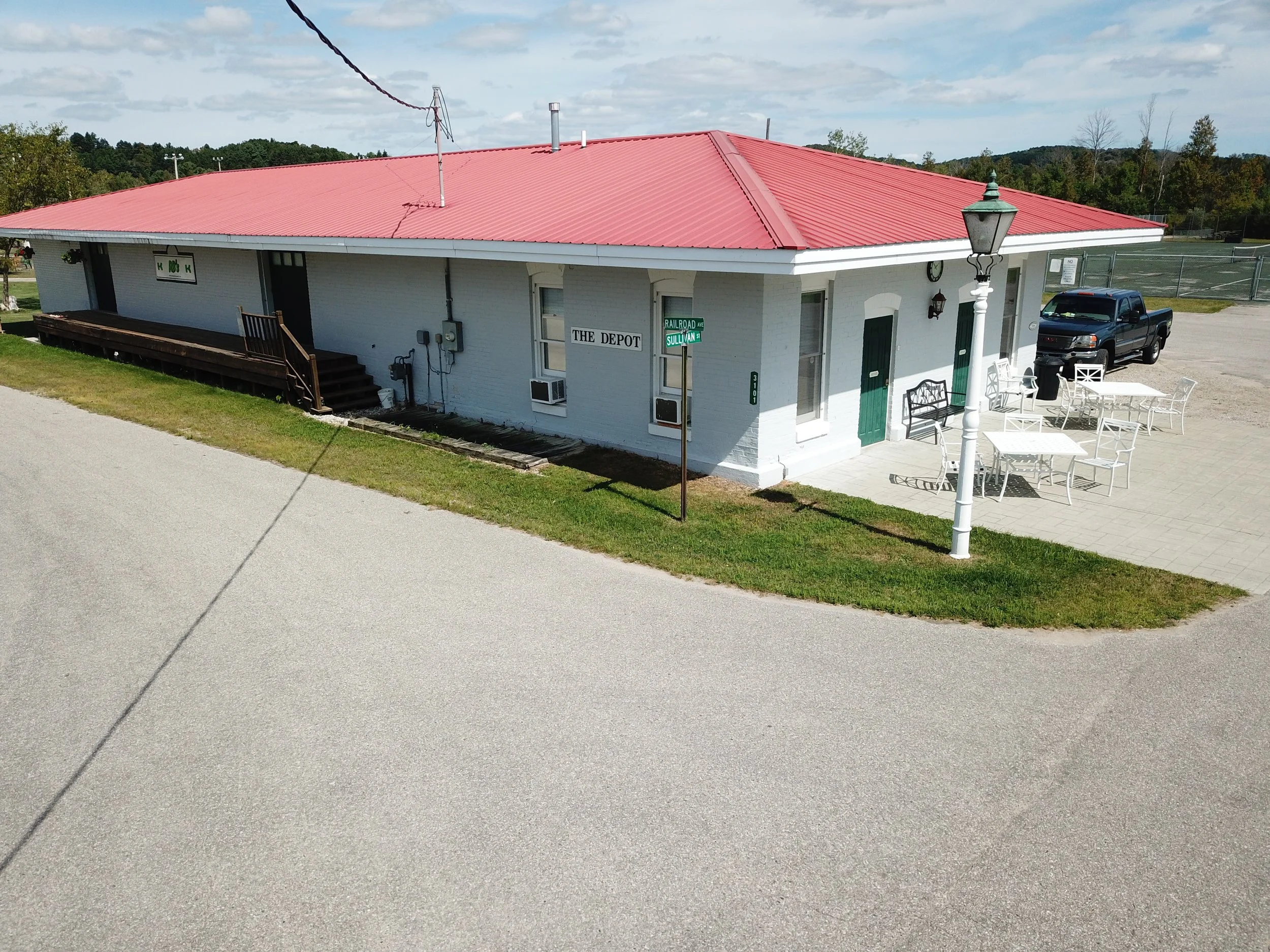 A white brick building with a red metal roof, a lamp post, outdoor seating, and a black pickup truck parked outside on a sunny day with scattered clouds.