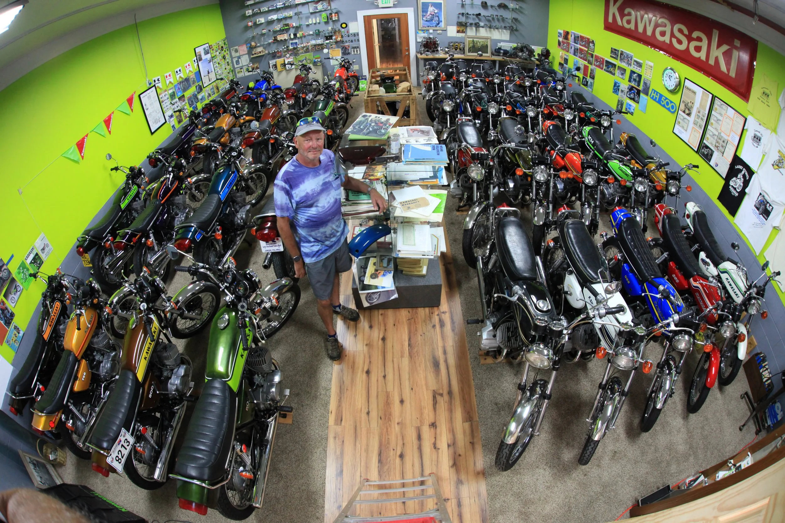 An indoor motorcycle showroom with a variety of Kawasaki motorcycles displayed in rows along the walls and center. A man is standing at a desk covered with books and papers, surrounded by more motorcycle models. The room has vibrant green and gray walls decorated with posters, photos, and signage related to Kawasaki motorcycles.
