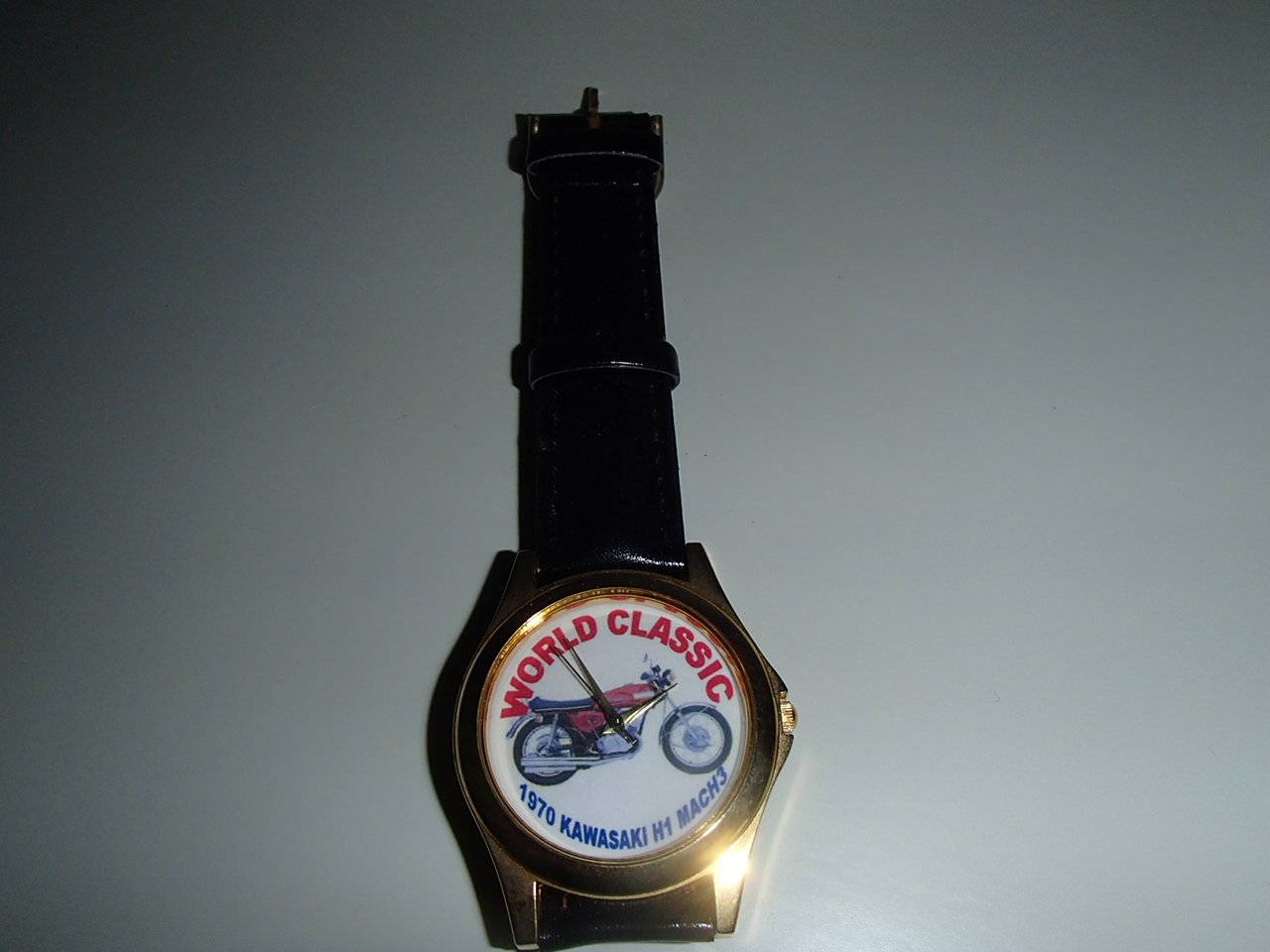 A wristwatch with a round face showing a motorcycle illustration and text 'World Classic 1970 Kawasaki H1 Mach 3' and a black leather strap.