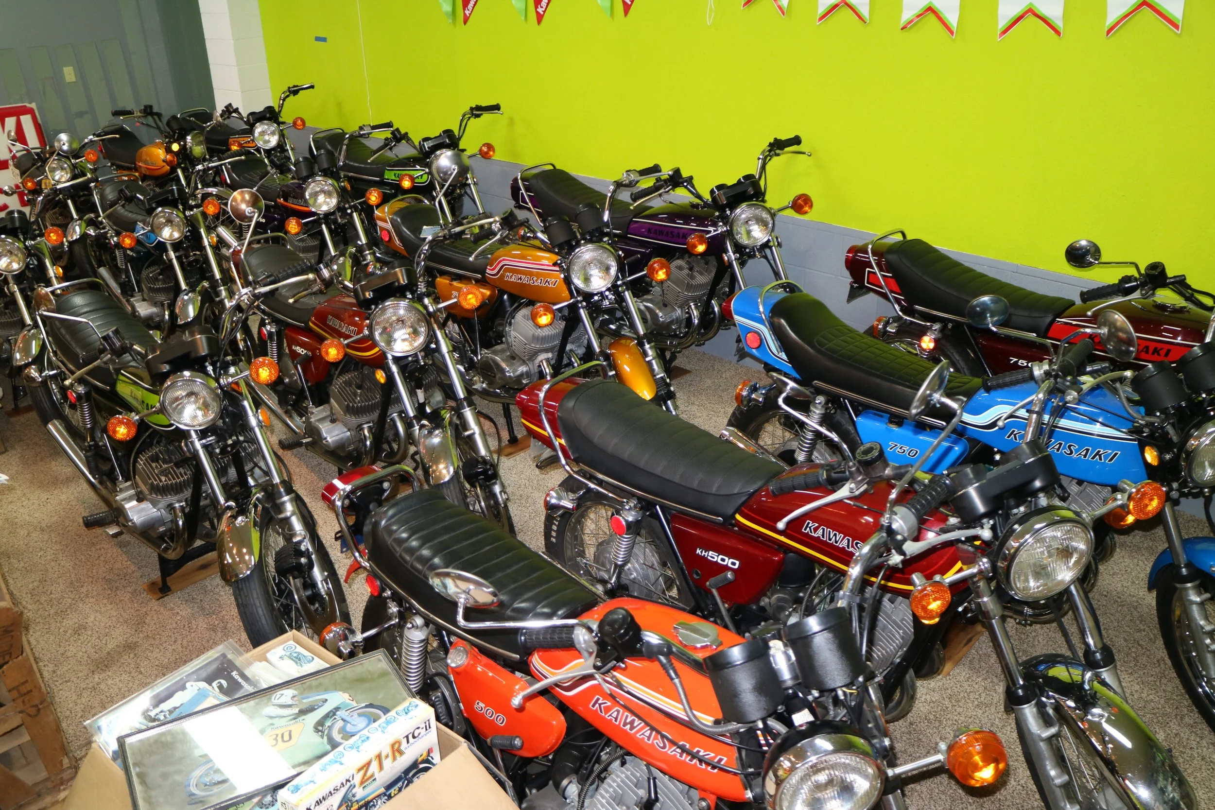 A collection of vintage Kawasaki motorcycles in various colors parked indoors against a bright lime green wall, with some displayed on wooden stands and others lined up on the floor.