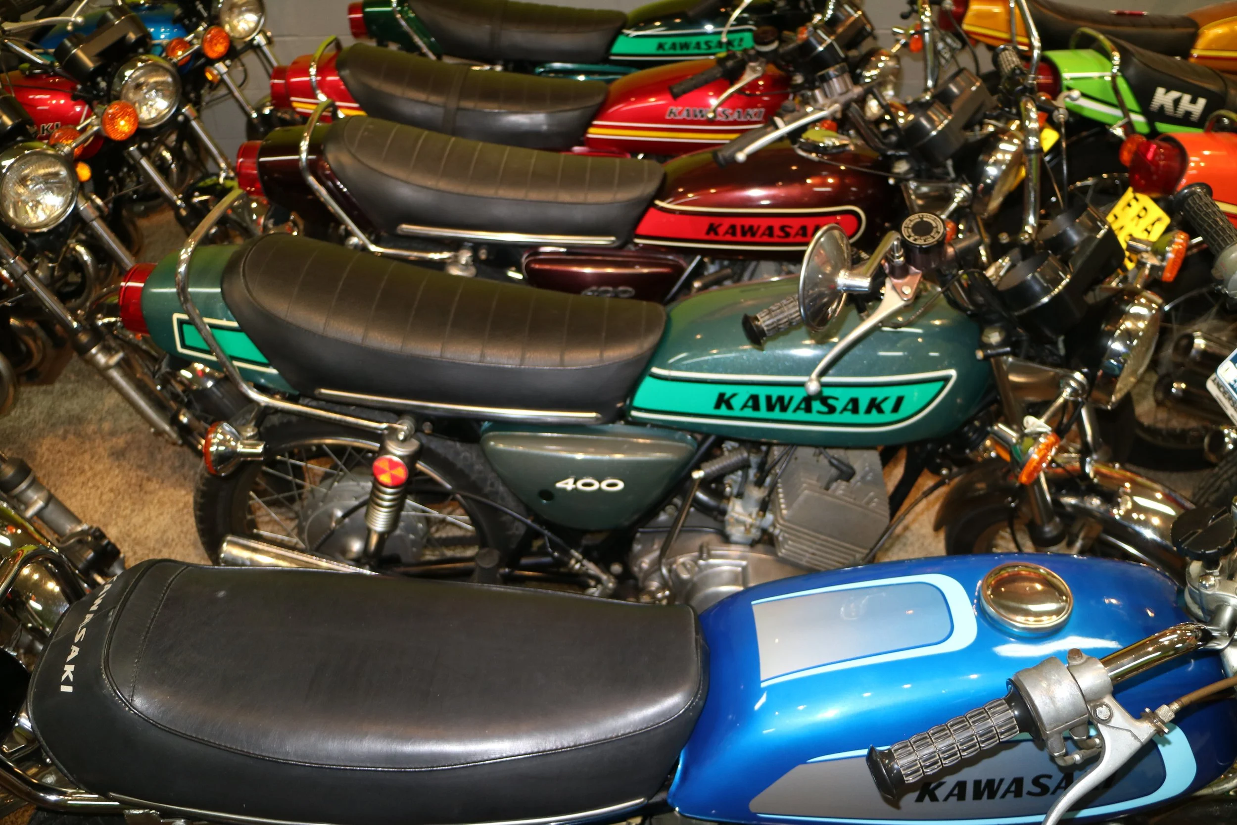 Multiple Kawasaki motorcycles parked closely together, featuring various colors including green, red, yellow, and blue, with visible seats, handlebars, and headlamps.