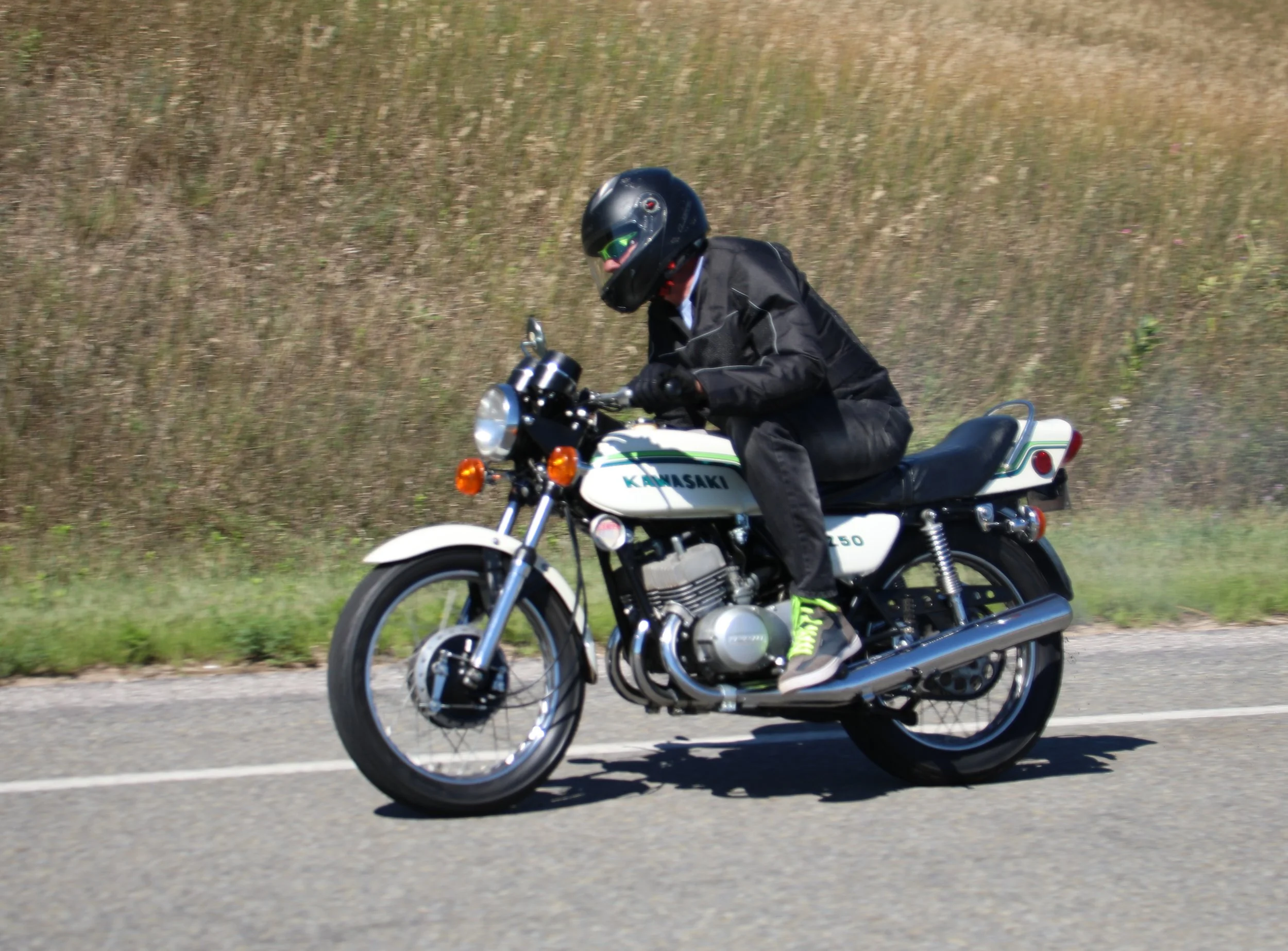 A person wearing a black helmet and jacket riding a Kawasaki motorcycle on a road with grass and a hill in the background.