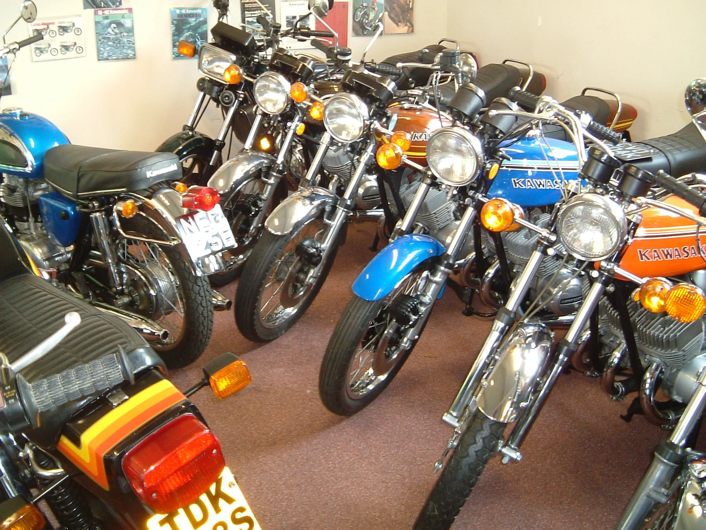 Multiple vintage Kawasaki motorcycles on display inside a showroom with posters on the wall.