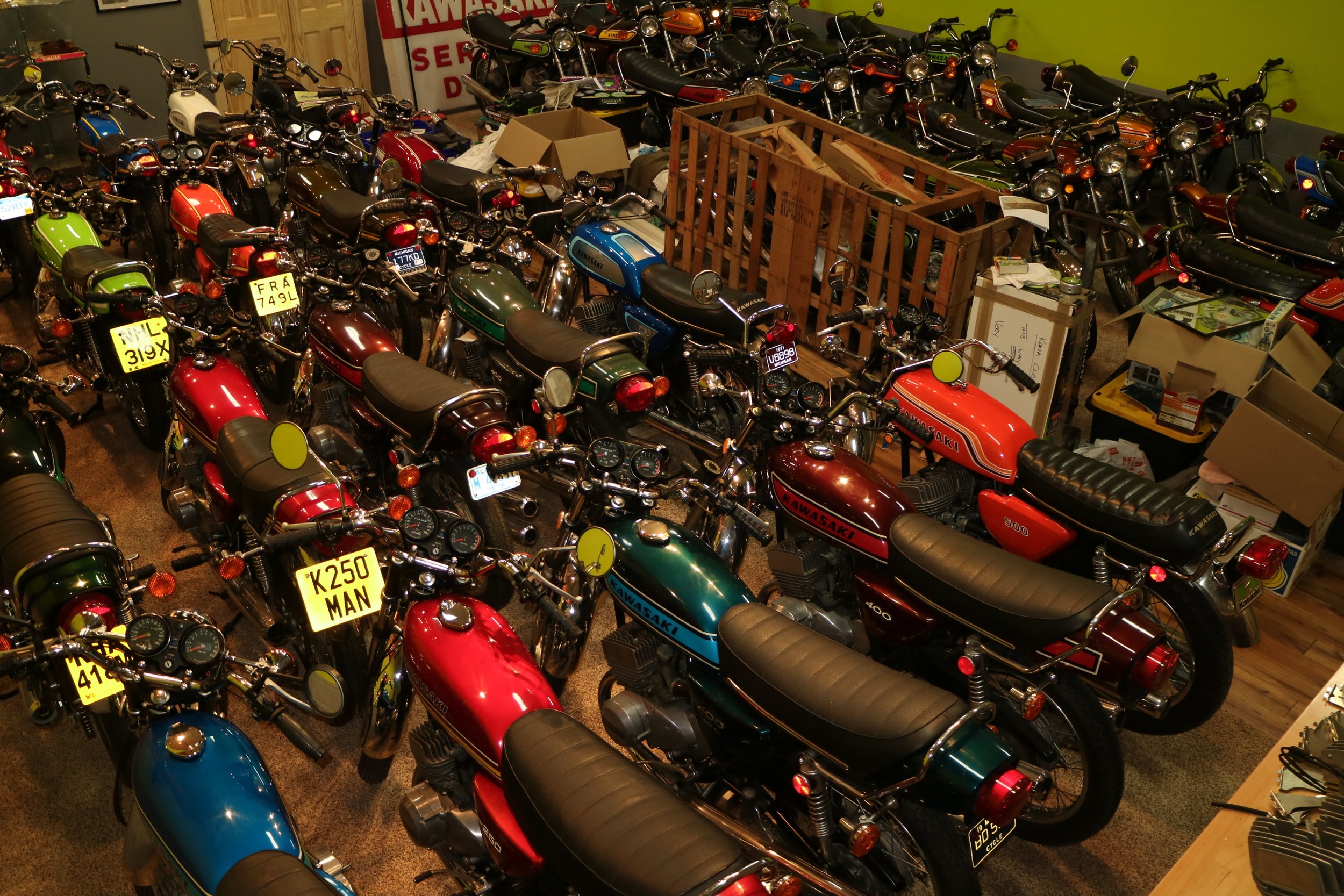 A large vintage motorcycle collection inside a storage or showroom area. The motorcycles are mainly Kawasaki, with various colors including red, green, blue, white, and black. Some motorcycles have license plates, and there are cardboard boxes and wo