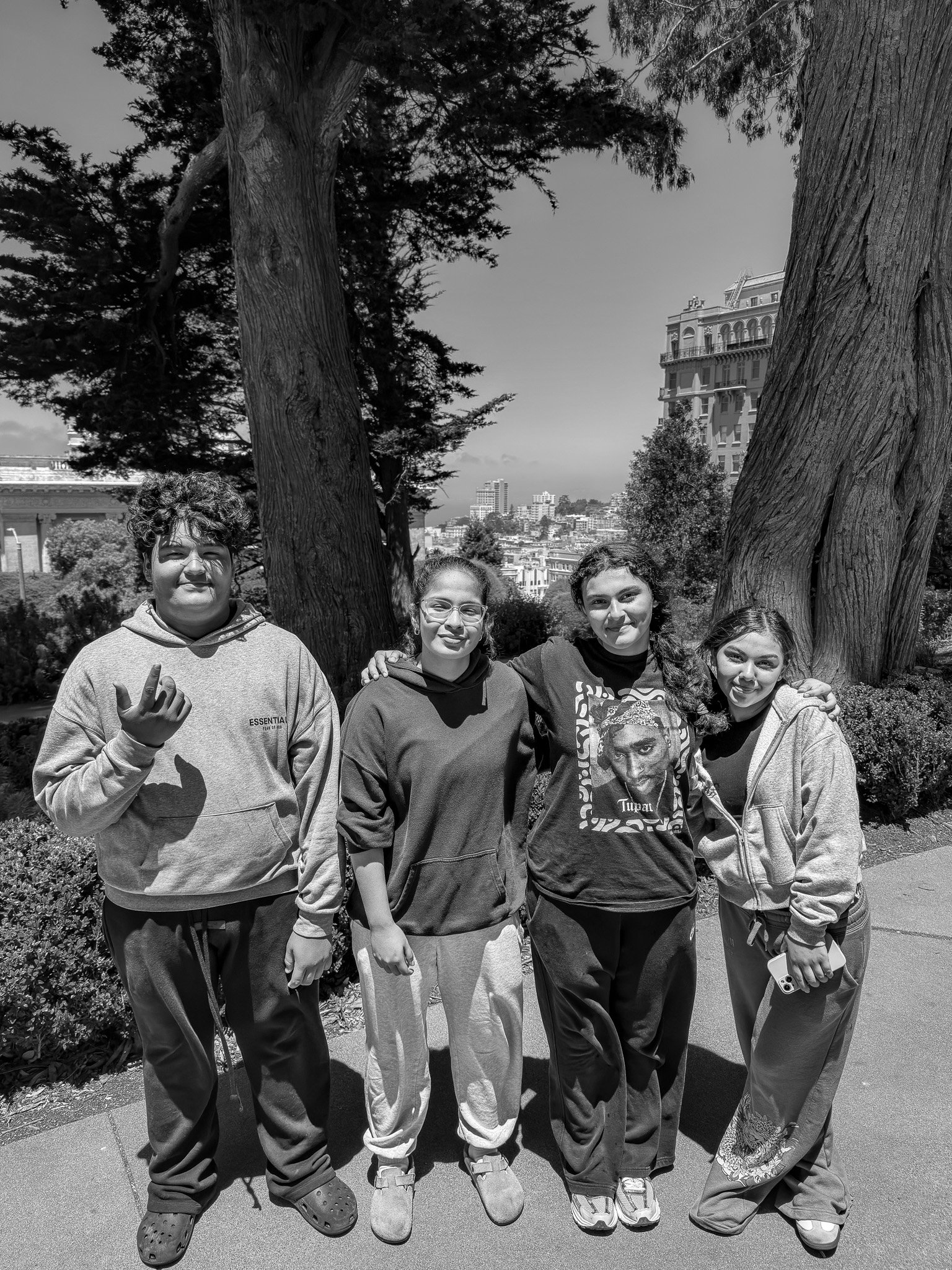 A group of five young girls standing together outdoors in front of trees and a city skyline, smiling and posing for the camera.