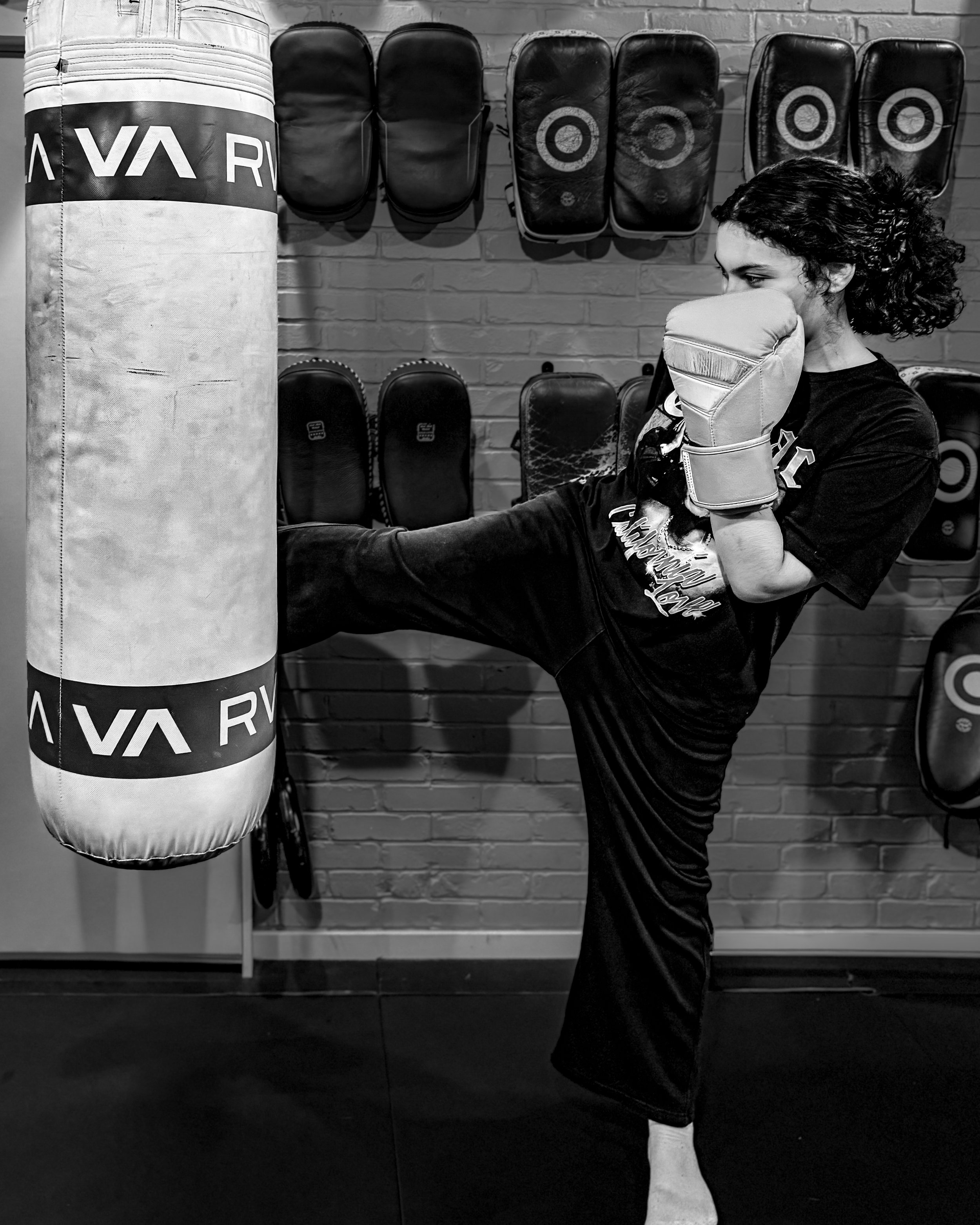 A woman wearing boxing gloves practicing a punch on a punching bag in a gym. The gym has a brick wall with multiple punch pads and chairs hung on the wall.