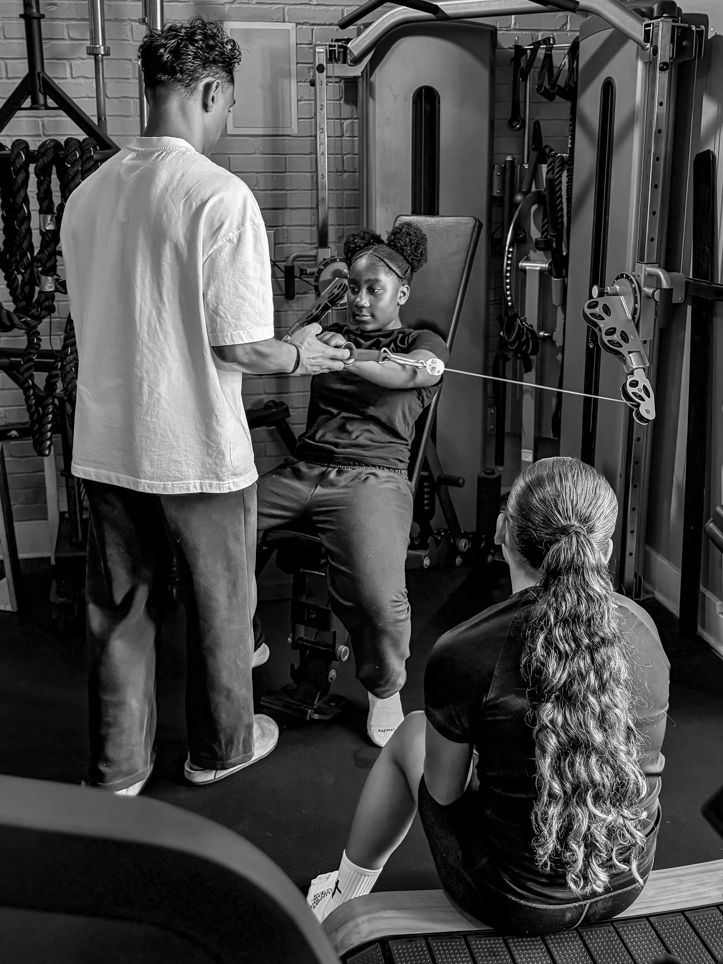 A girl with curly hair sitting on a bench in a gym, receiving physical therapy or exercise instructions from a trainer. The trainer is assisting her with a resistance exercise using a pulley system. Another woman is sitting on the floor, observing the session.